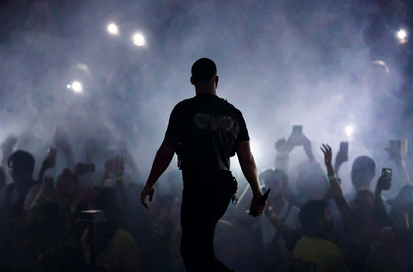 Drake 桌布 壁紙 — 表演者的剪影穿過煙霧繚繞的舞台燈光，充滿活力的觀眾在黑暗中舉起手機。