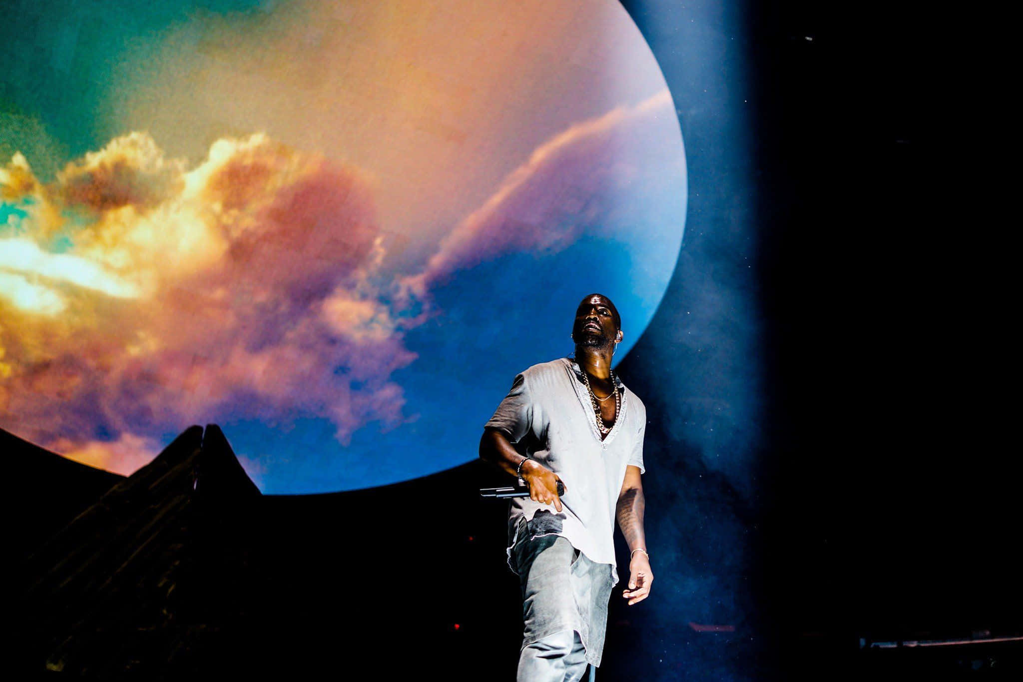 Kanye 桌布 壁紙 — 表演者站在舞台上，上方是巨大的投影天空，有鮮豔的雲朵和發光的星球。