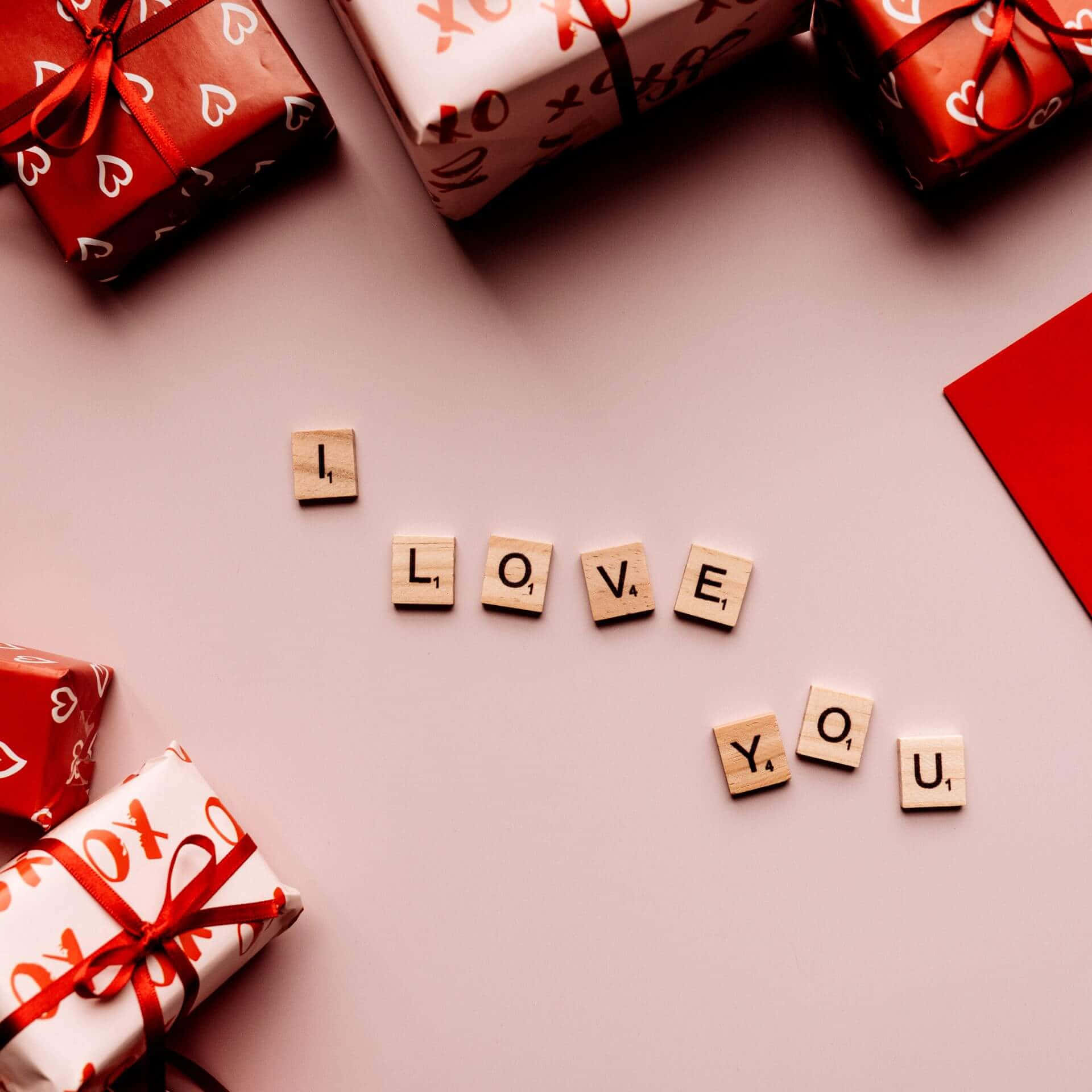 表達愛意的美好方式 壁紙 — 木製拼字磚拼出「I LOVE YOU」，周圍係紅白心形圖案禮物，放喺柔軟粉紅色表面上。