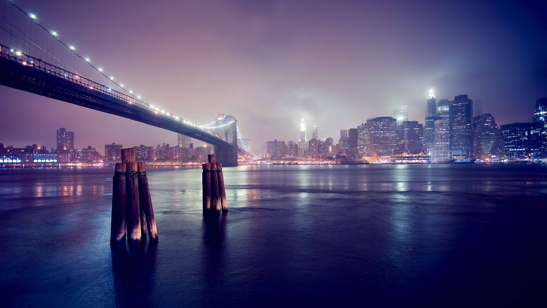 夜晚在水面上亮燈的布魯克林大橋，背景是城市天際線。