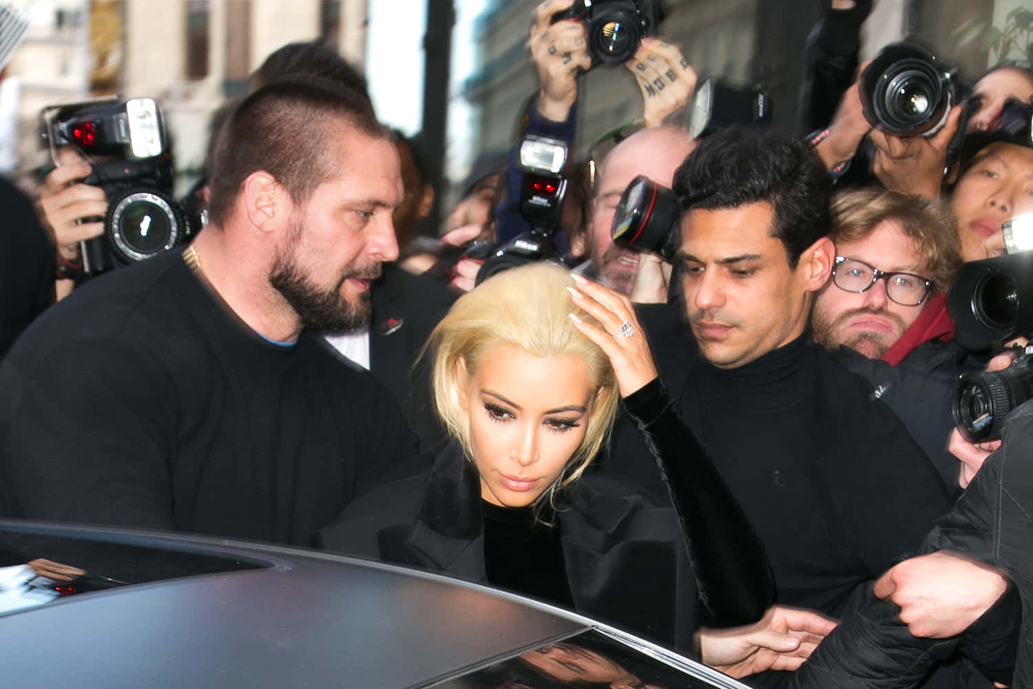 Kim Kardashian 由兩名男子護送，穿過一群攝影師，靠近一輛汽車。