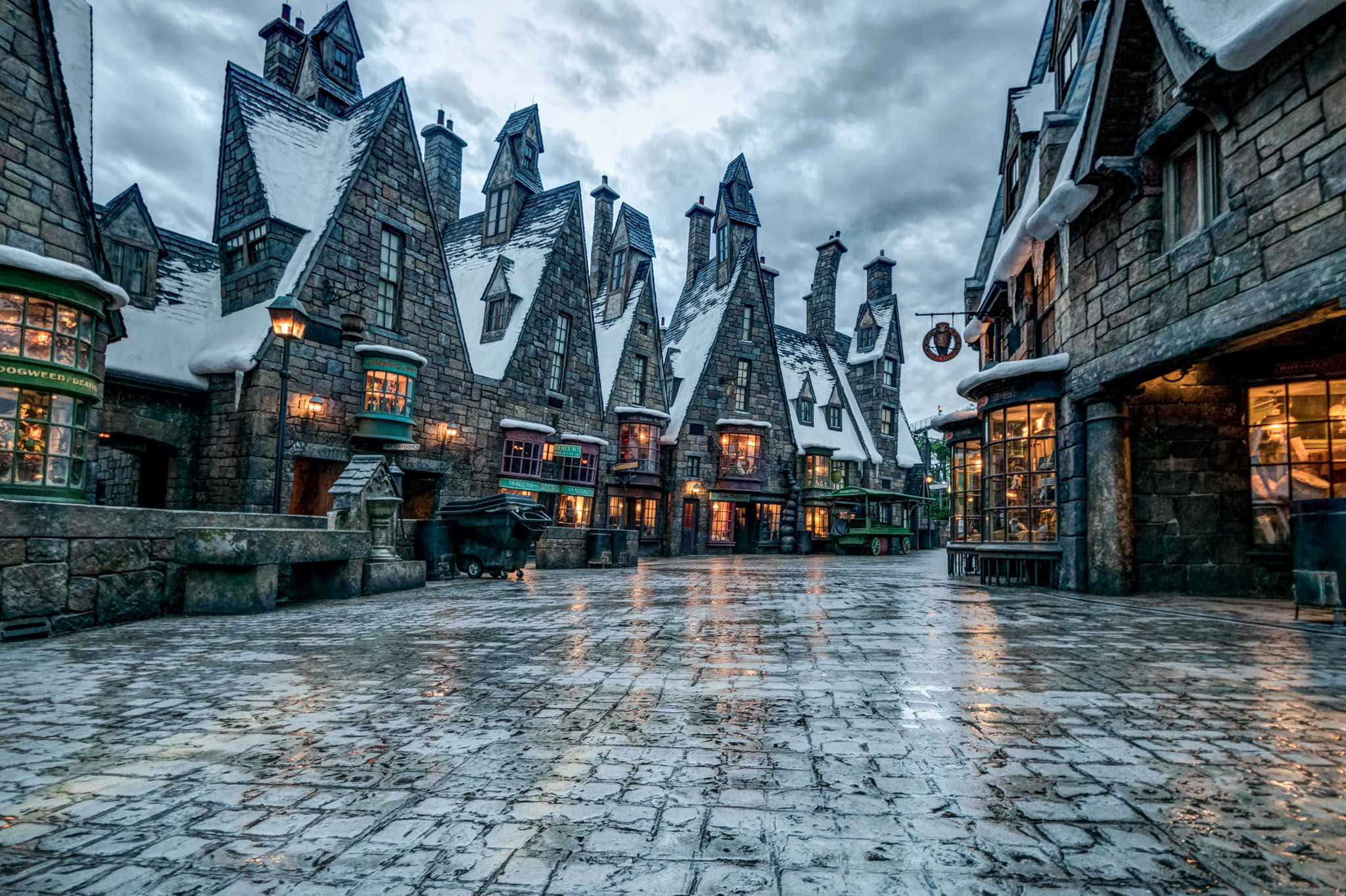 Wizarding World 桌布 壁紙 — 被雪覆蓋的中世紀風格建築排列在一條濕漉漉的鵝卵石街道上，陰沉的灰色天空下，溫暖的窗戶閃爍著光芒。