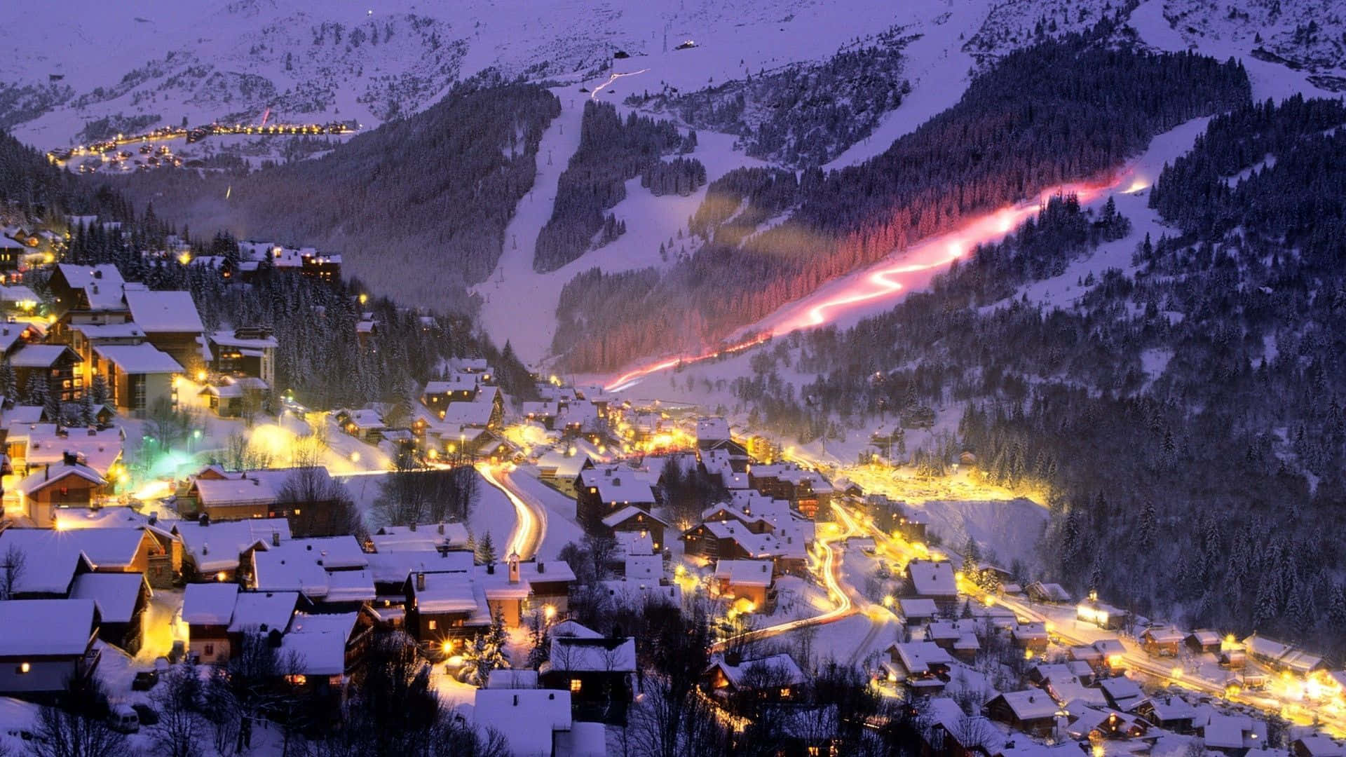Snowy Village 桌布 壁紙 — 被雪覆蓋的山村在紫色暮色天空下閃爍著溫暖燈光，並有照明滑雪道。