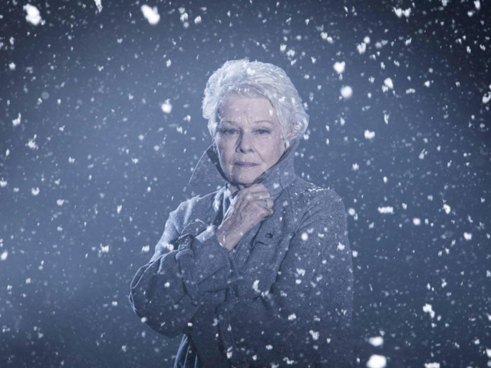 裘蒂·丹奇雪中風情 壁紙 — 一位白髮老婦人穿著厚重外套，在寒冷的藍色燈光場景中，雪花在她周圍旋轉，她堅定地站立著。