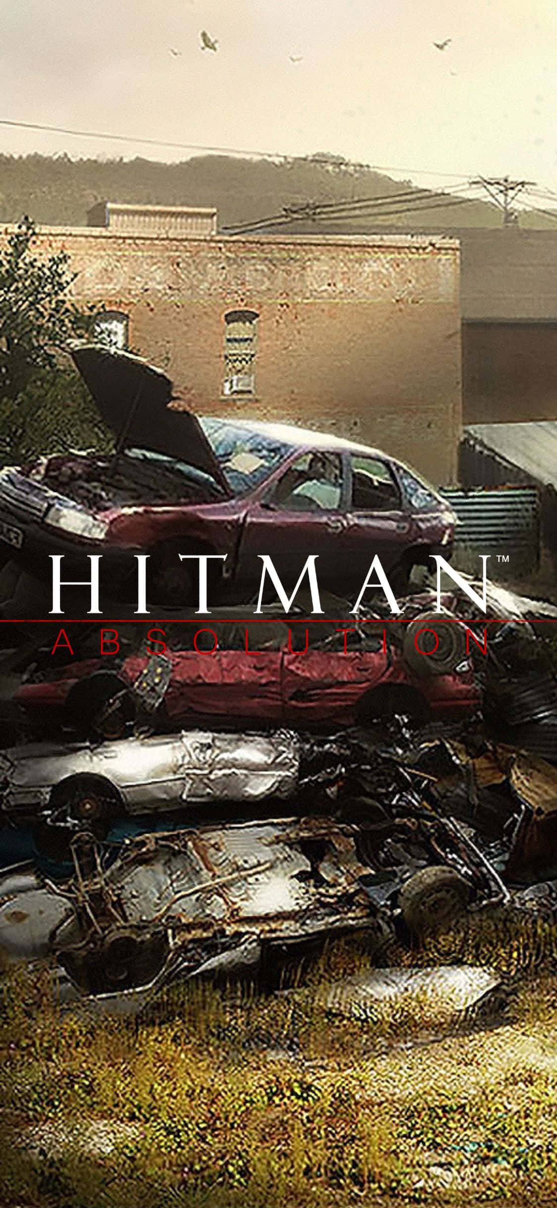 裝備上陣，與《Hitman》展開終極任務 壁紙 — 一堆報廢汽車，其中一輛引擎蓋打開，停放在磚砌建築旁的草地上，天空朦朧。