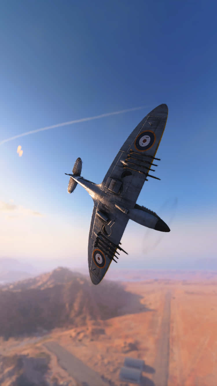 裝備導彈的 Supermarine Spitfire 下方 Android 飛機背景 壁紙 — 一架復古英國戰鬥機在晴朗的藍天中翱翔，下方是陽光照耀的沙漠景觀，遠處有山脈。