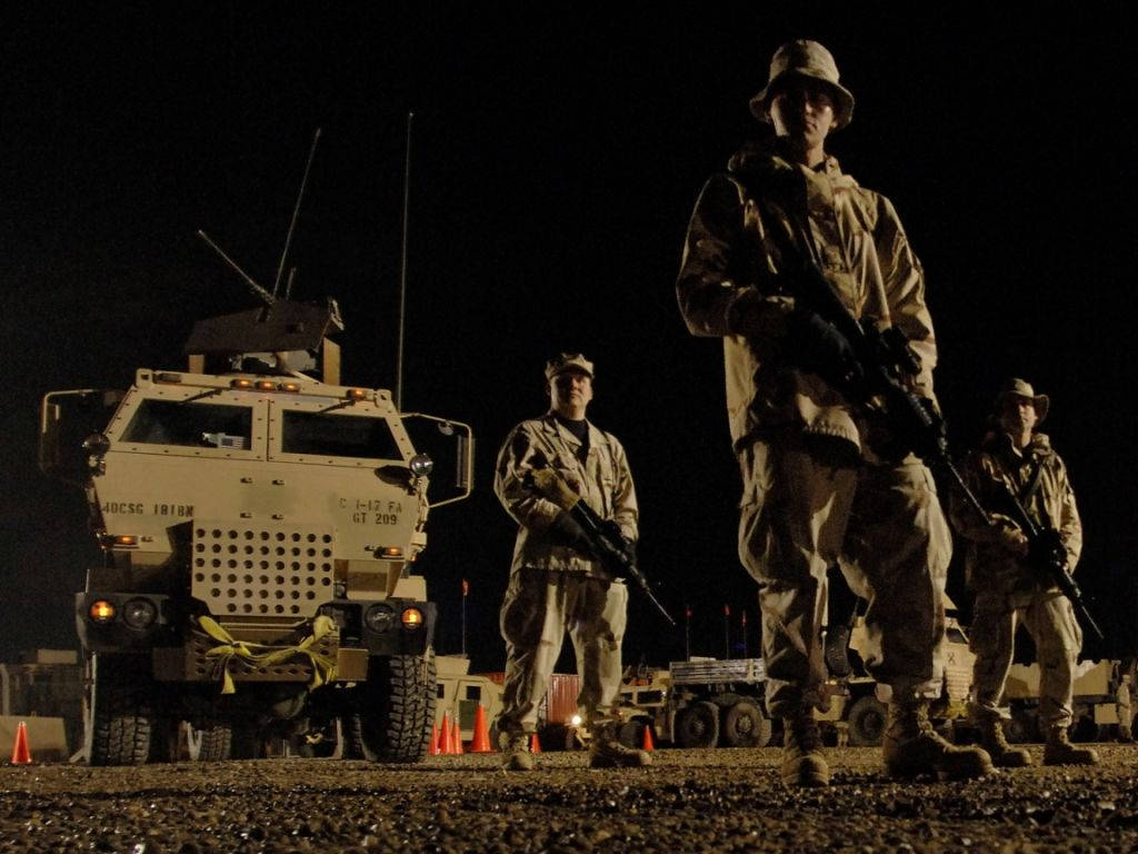 夜間場景，士兵持步槍在沙漠地形中的軍用車輛附近。
