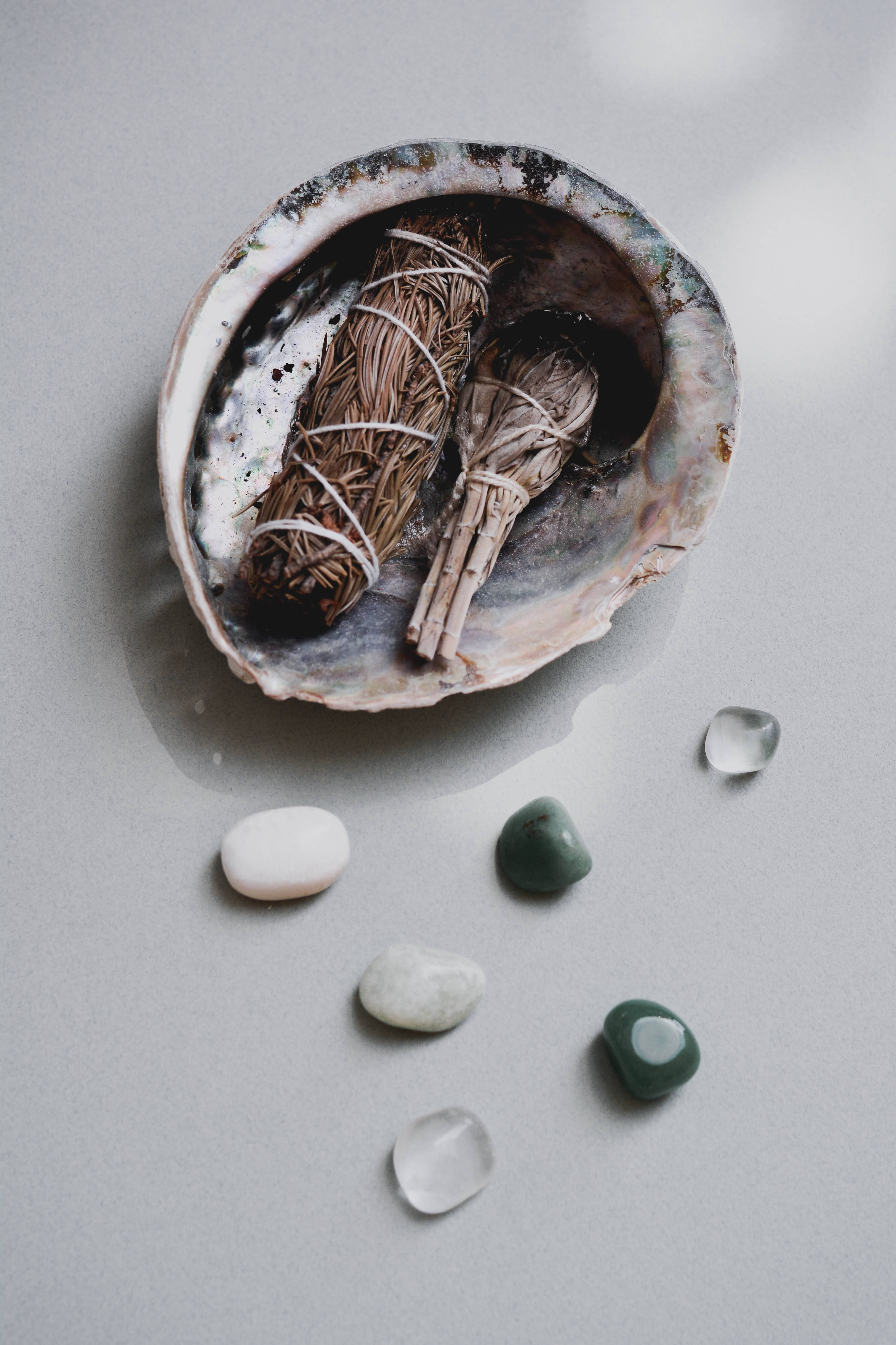 裝飾著療癒水晶與 sage 的神祕鰈貝殼 壁紙 — 兩支鼠尾草煙燻棒放在一個虹彩鮑魚殼內，周圍是光滑的白色和綠色石頭，置於柔軟的灰色表面上。