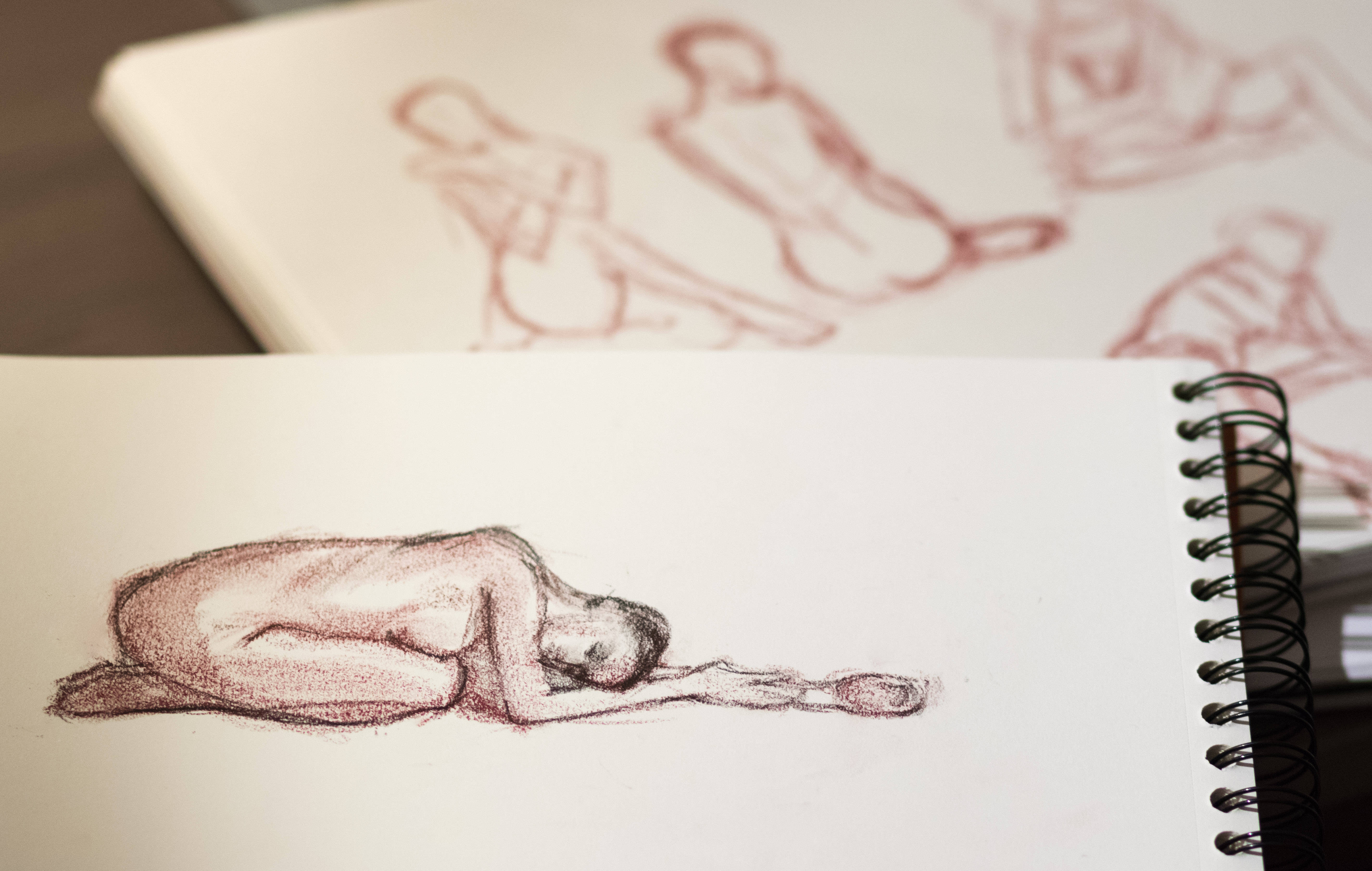 裸體人體鉛筆素描 壁紙 — 在螺旋裝訂素描本頁面上，一幅詳細的紅色粉筆素描，描繪一個胎兒姿勢的裸體人物。