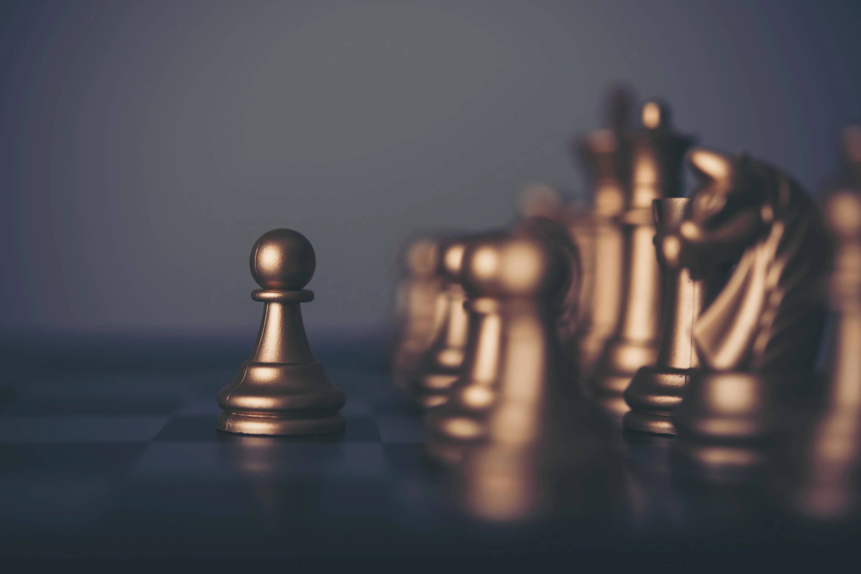褪色銅色兵棋 壁紙 — 一個孤獨的金色兵卒在棋盤上清晰對焦，而其他棋子則在柔和燈光下模糊成背景。