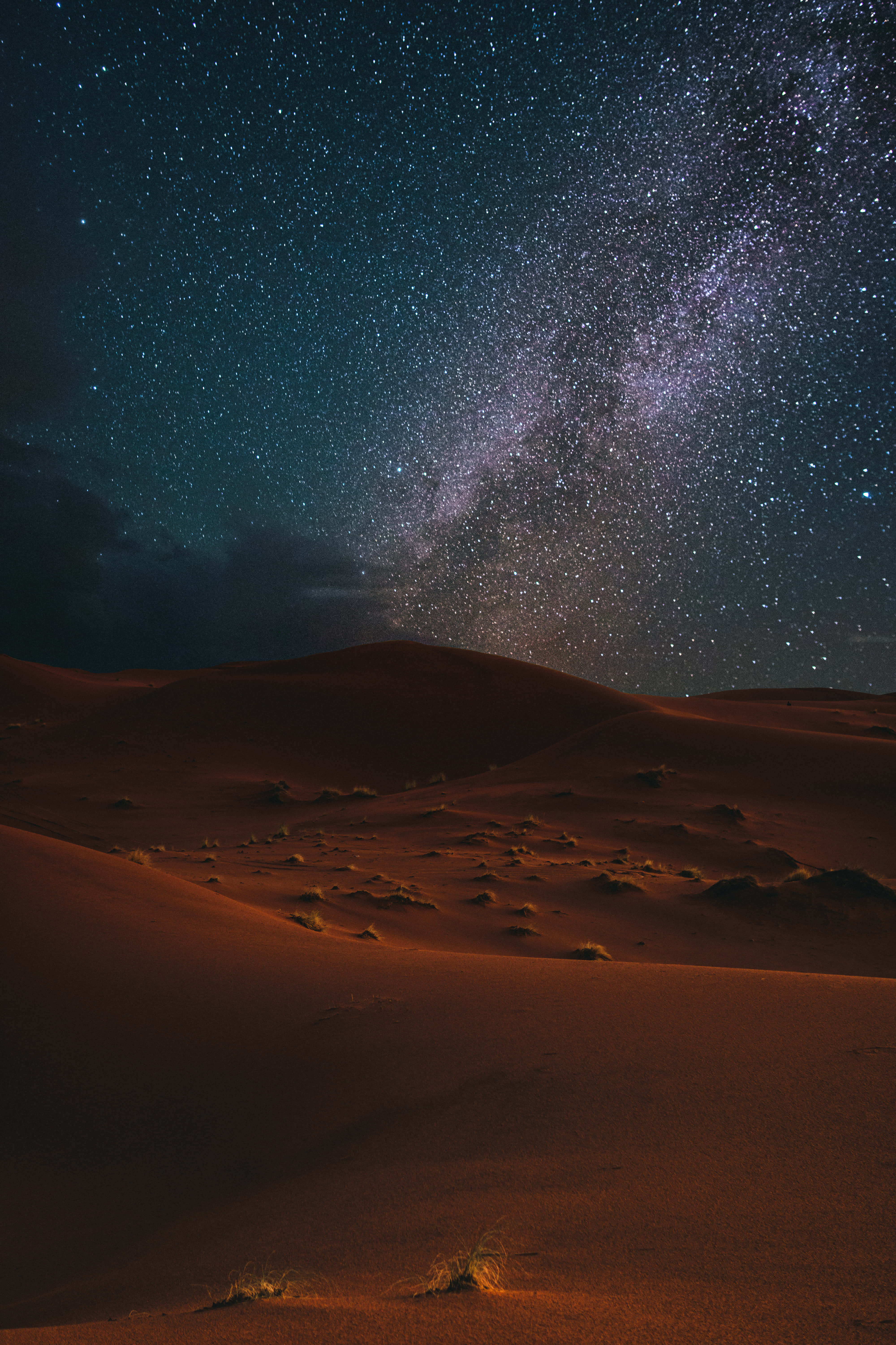 西南沙漠中的雄偉山脈 壁紙 — 繁星點點的夜空，銀河在深藍色廣闊天空下拱形掠過發光的橙色沙漠沙丘。