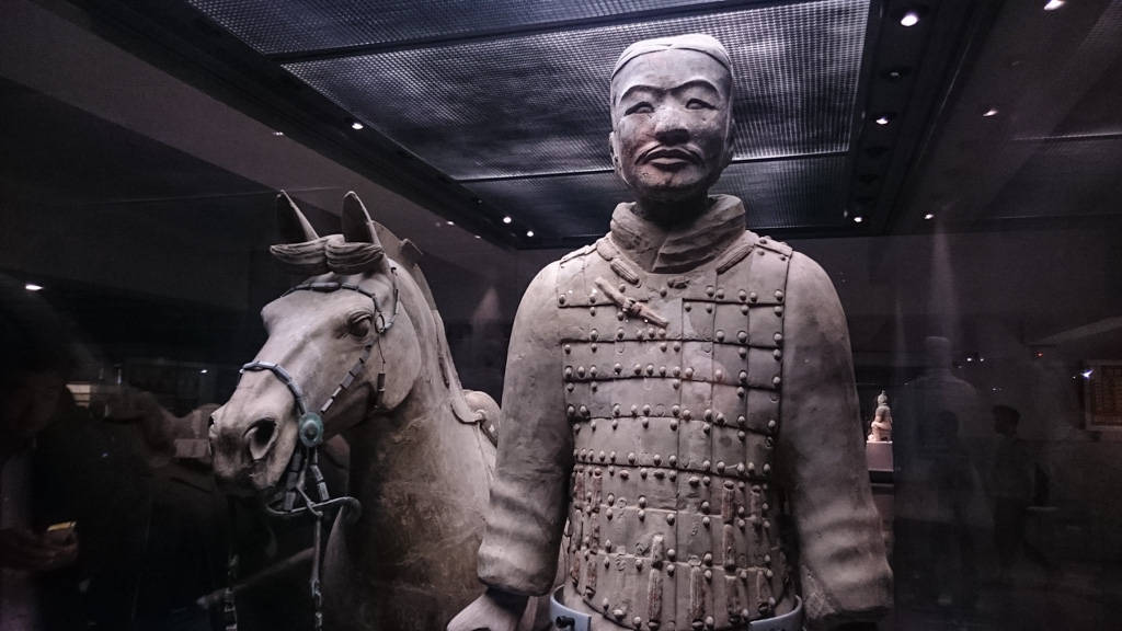 西安中國騎馬武士雕像 壁紙 — 兵馬俑及馬匹雕像在博物館燈光下展出，背景隱約可見參觀者。