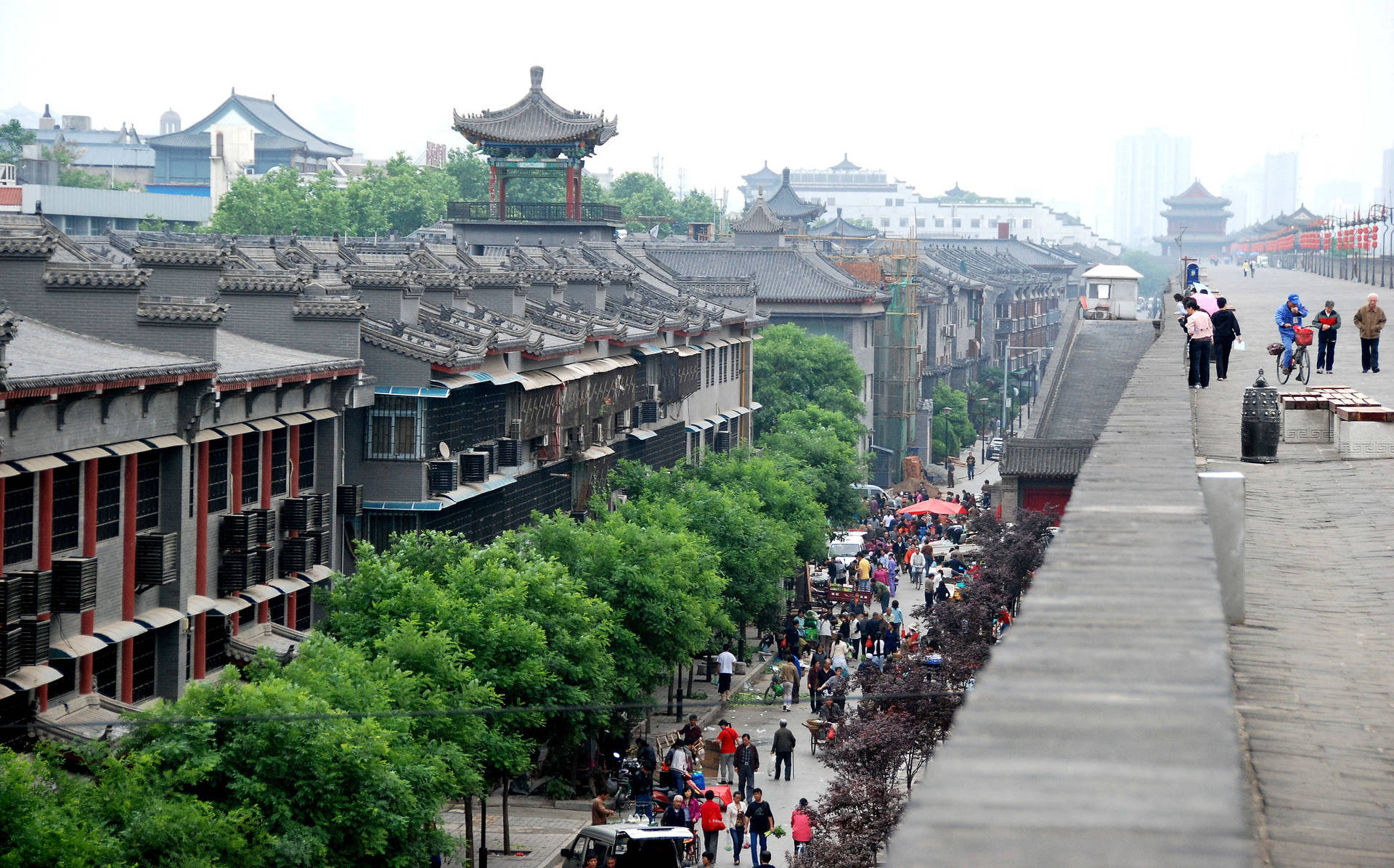 西安城牆的落差 壁紙 — 一條熱鬧的傳統中國街道，有瓦片屋頂、綠樹，以及行人在陰天沿著石坡行走。