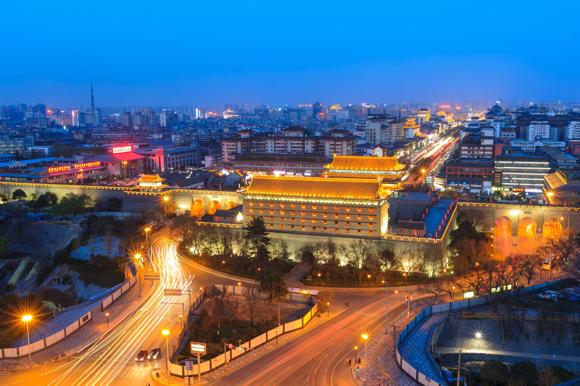 西安夜景燈光 壁紙 — 古代中國城牆的鳥瞰圖，在黃昏時溫暖地發光，背景有汽車燈光軌跡和現代建築。