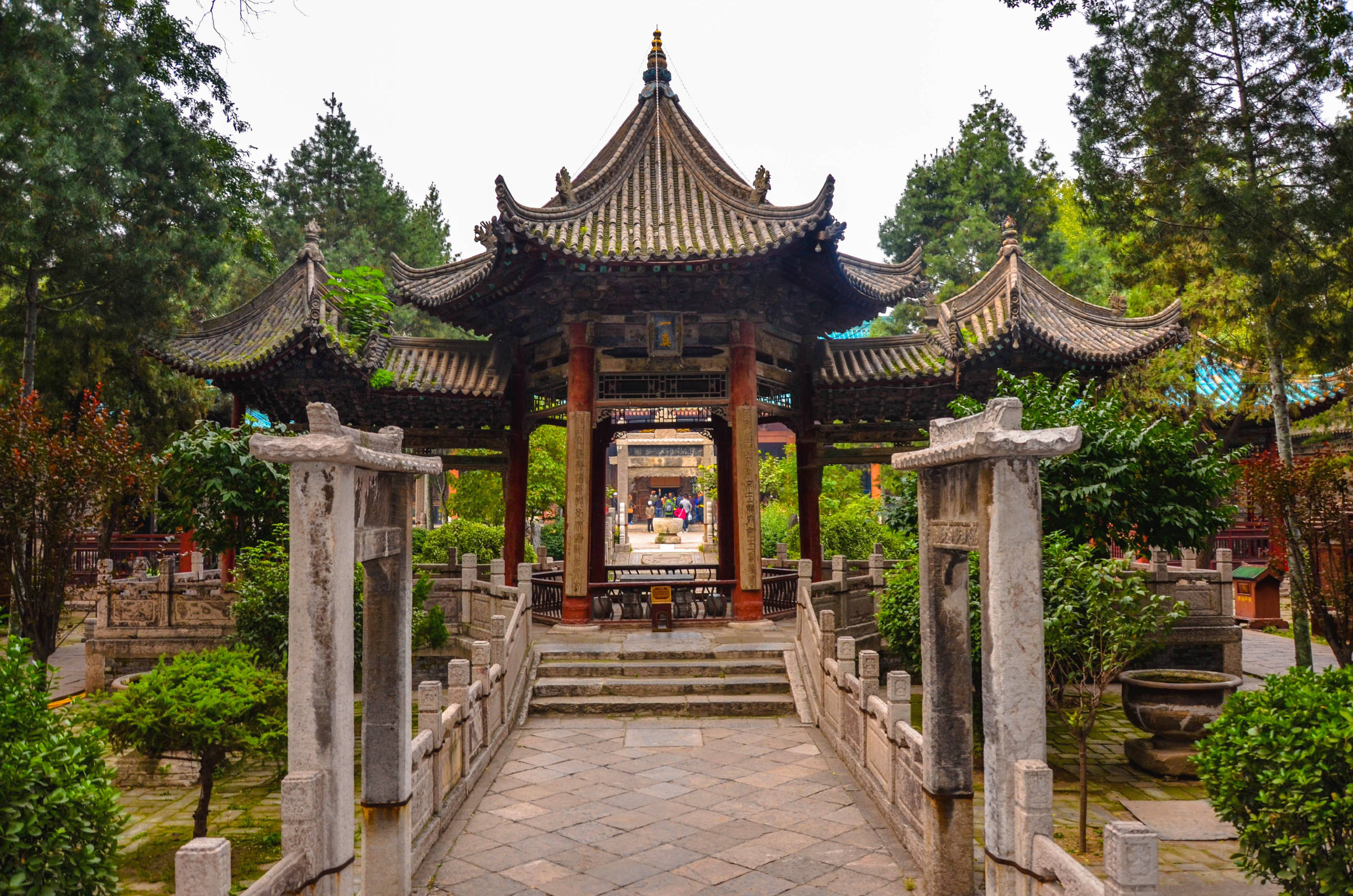 西安大清真寺 壁紙 — 一座傳統中式亭閣，擁有彎曲的瓦片屋頂，矗立在石橋盡頭，周圍環繞著茂密的綠樹和灌木。
