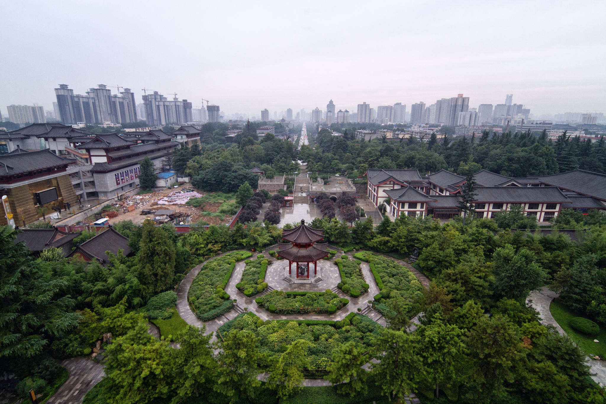 西安大清真寺鳥瞰圖 壁紙 — 傳統中式亭台樓閣和花園的鳥瞰圖，周圍環繞著茂密的綠植，遠處朦朧中可見現代城市天際線。