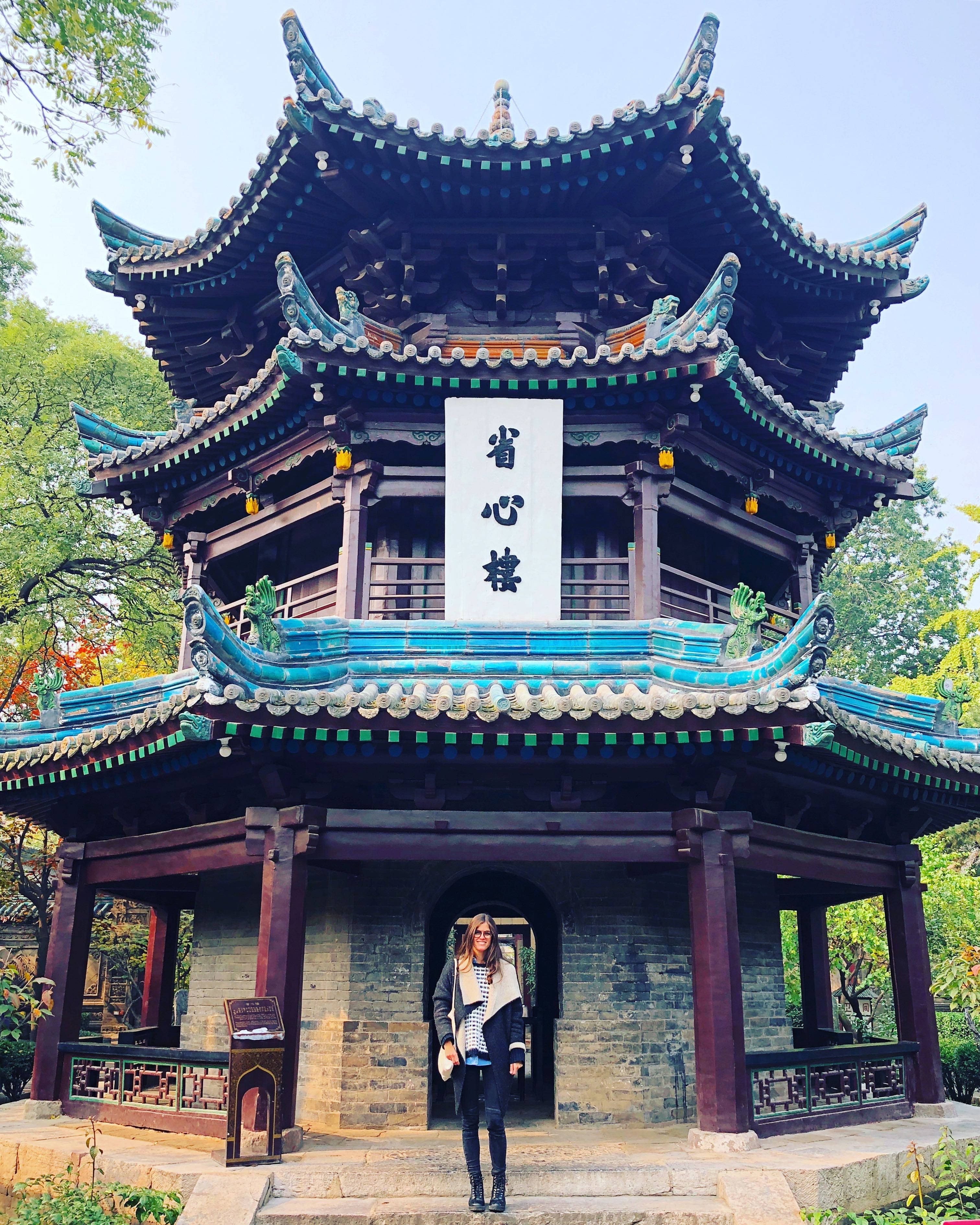 西安清真寺前的女子 壁紙 — 一位女士站在傳統中式亭閣前，亭閣有彎曲的藍綠色瓦頂和木柱，周圍是茂密的綠樹，天空晴朗。