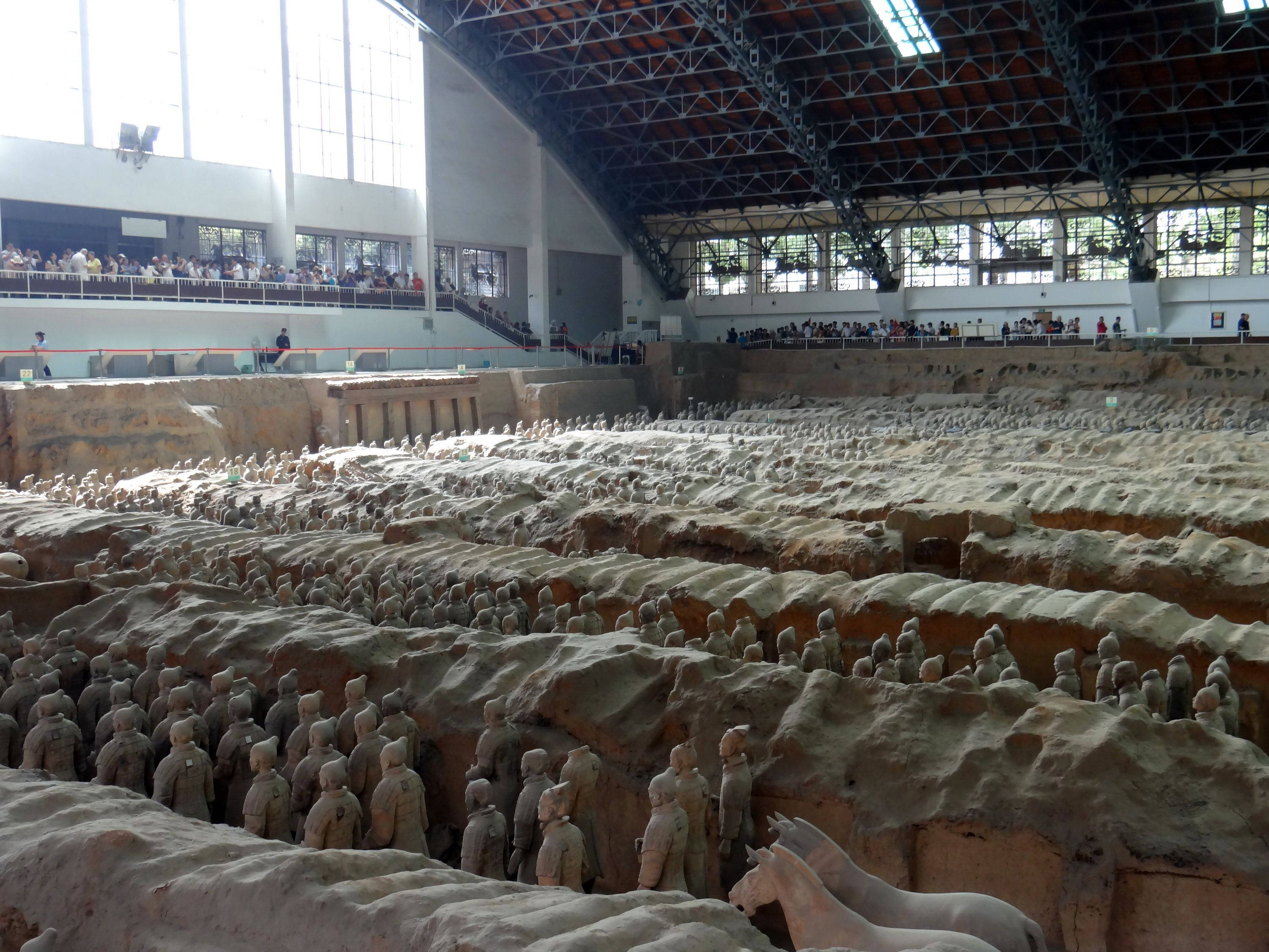 西安無數石像 壁紙 — 一排排古老的兵馬俑站在現代展覽館下的土坑中，參觀者從陽台上觀看。