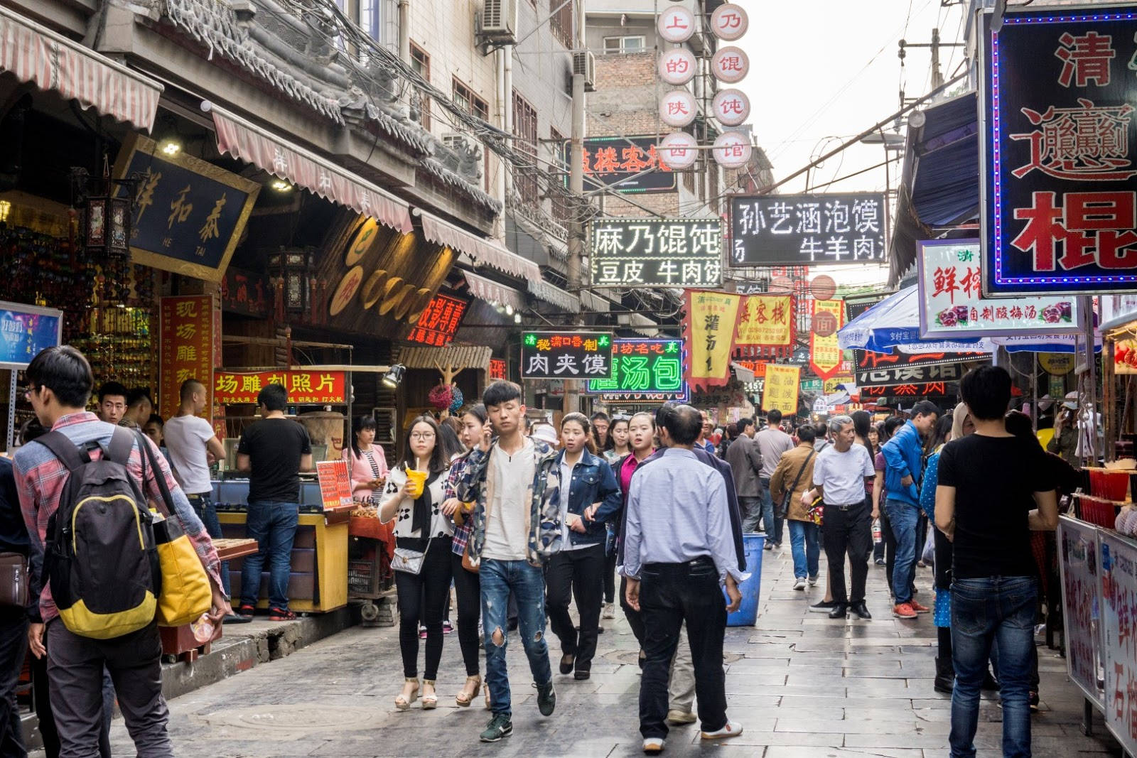 西安熱鬧街景 壁紙 — 亞洲城市中一條繁忙狹窄的街道，擠滿行人，頭頂懸掛著色彩繽紛的發光食物招牌。