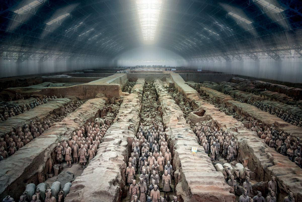 西安秦俑軍隊 壁紙 — 一排排古老的兵馬俑在考古坑中廣闊的工業屋頂下以精確的隊列站立。