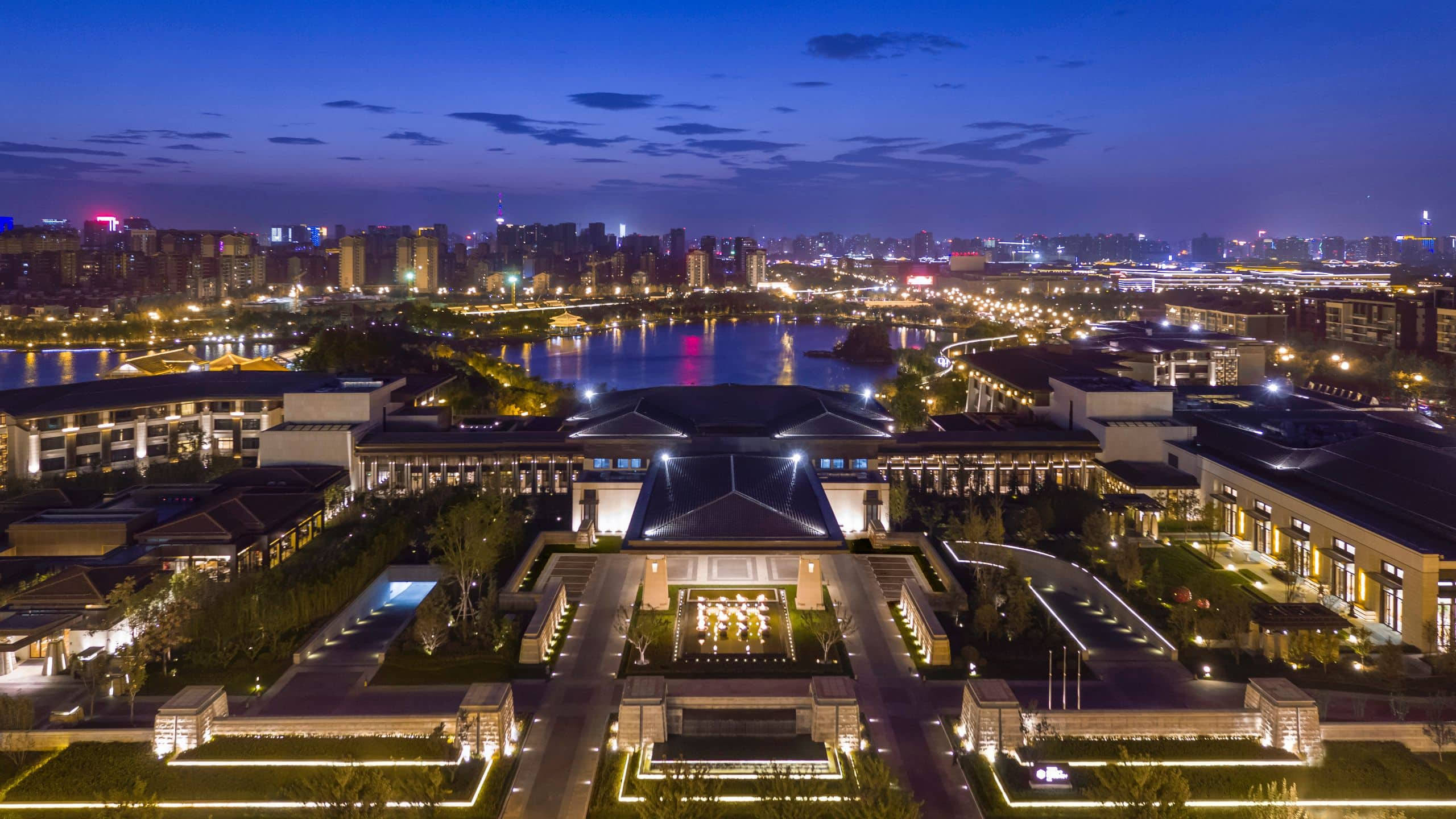西安豪華酒店夜景 壁紙 — 一座宏偉的現代建築群在黃昏時分被溫暖的燈光照亮，燈光倒映在平靜的河面上，周圍是廣闊的城市景觀。