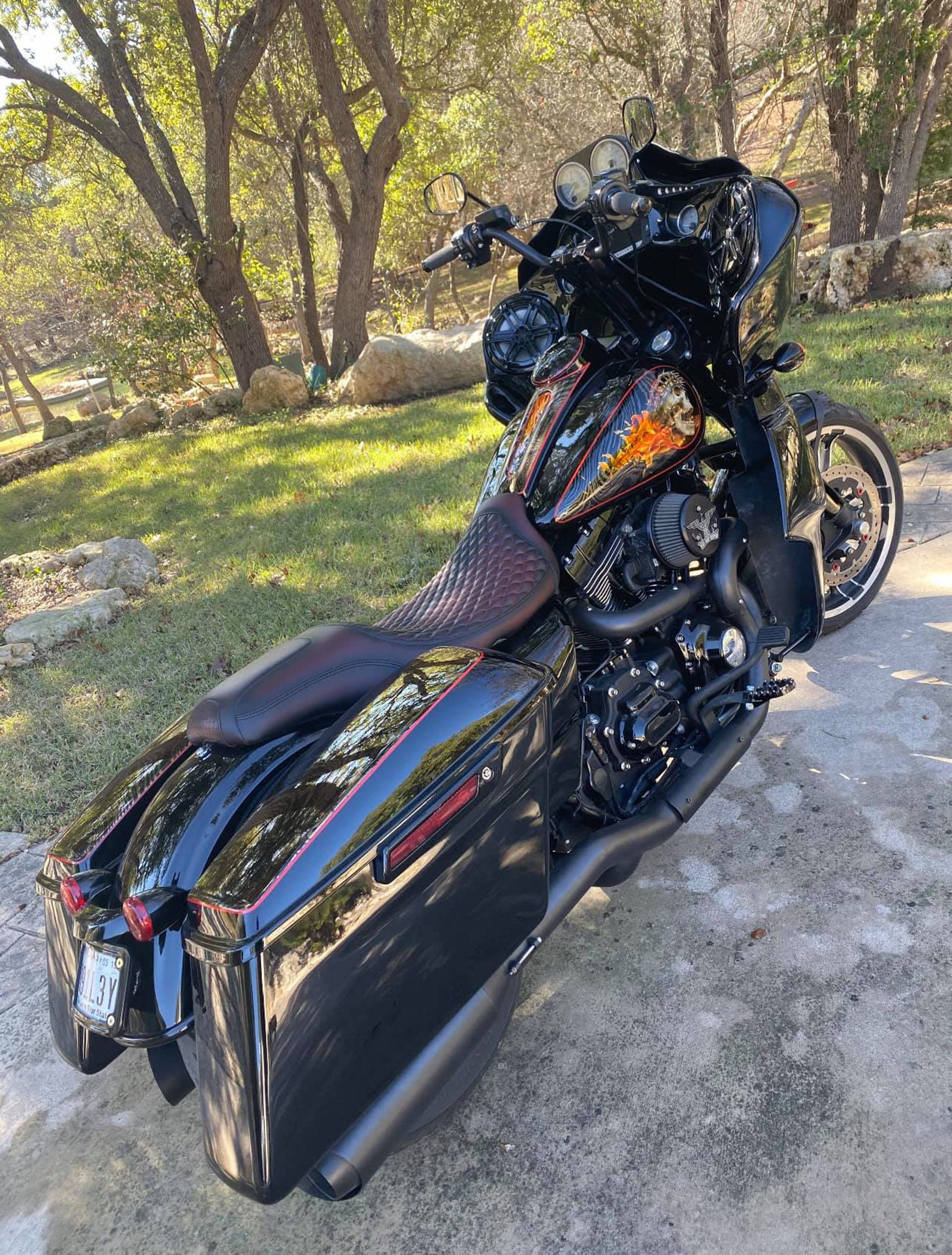 西海岸車廠改裝Bagger 壁紙 — 一架光澤黑色旅行電單車帶有自訂火焰貼紙，停放在混凝土墊上，周圍是陽光照射的樹木和草地。