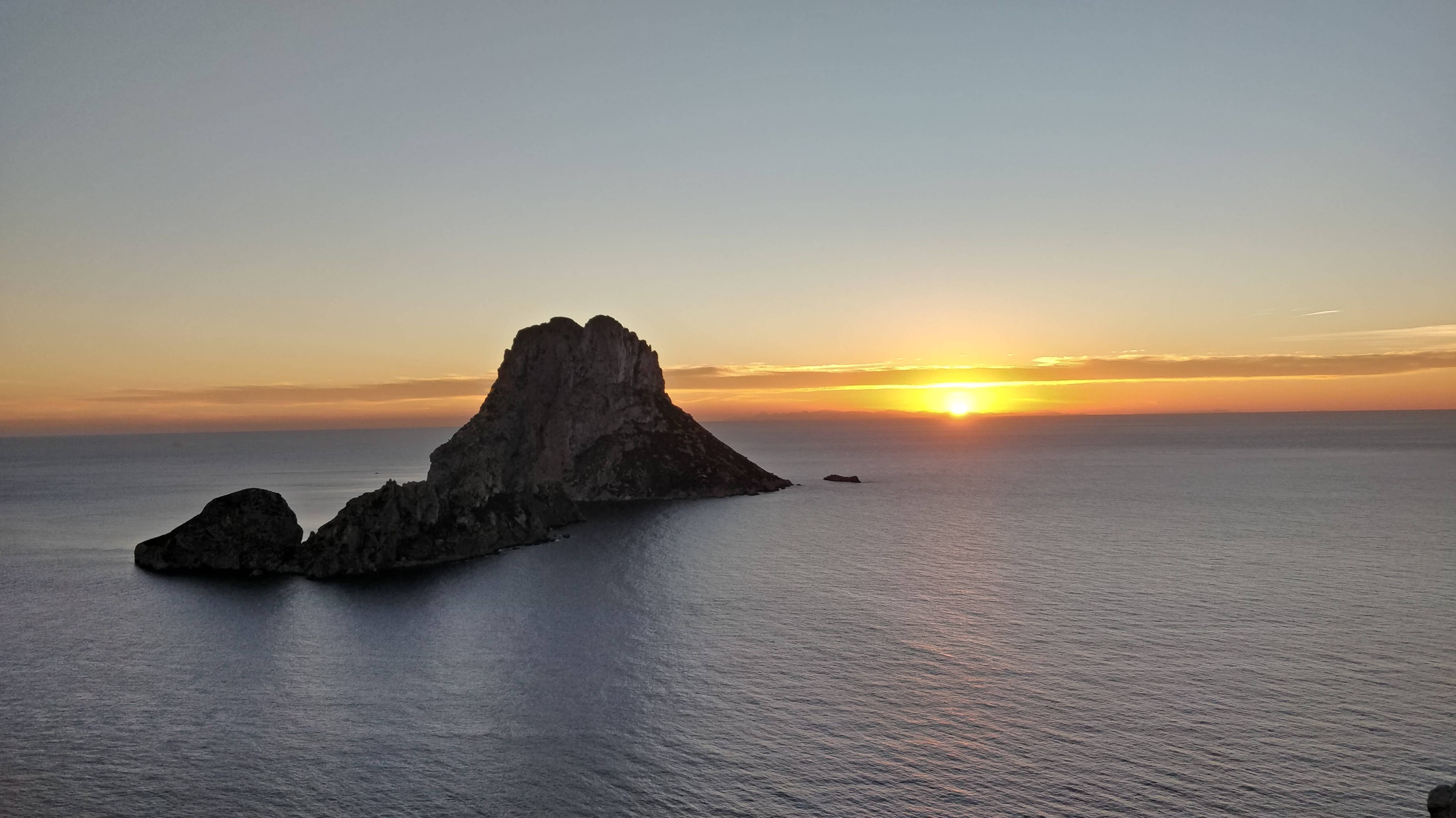 西班牙伊比薩島埃斯·韋德拉日落 壁紙 — 一個孤獨崎嶇的島嶼在平靜的海面上，襯著金色的日落形成剪影。