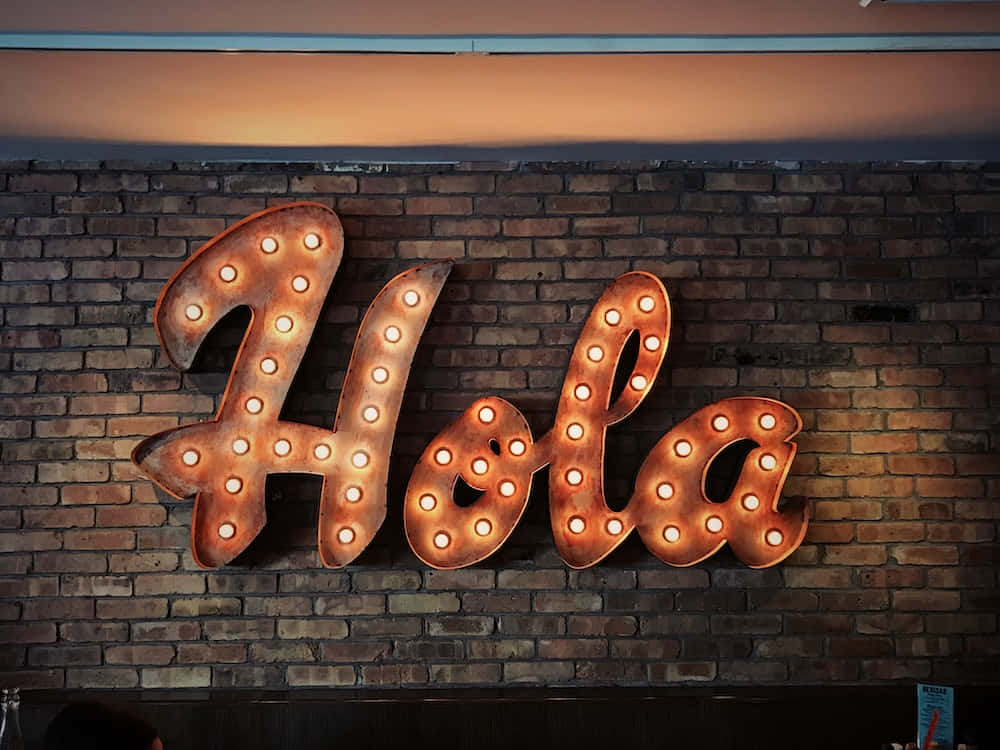 西班牙壯麗風景 壁紙 — 一個發光的招牌，用草書字母拼出「Hola」，帶有溫暖的燈泡，安裝在質樸的磚牆上。
