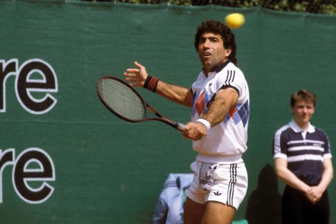 西班牙網球選手曼努艾爾·奧蘭特斯於1985年世界團體杯比賽 壁紙 — 一位身穿復古Adidas裝備的男網球手，在綠色背景下揮拍擊打黃色網球。