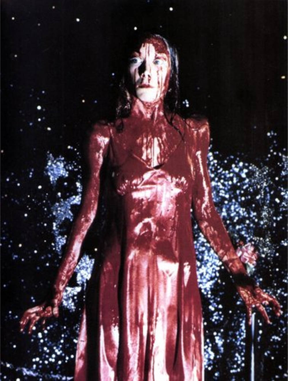 西茜·斯派克在《Carrie》中的經典場景 壁紙 — 一個身穿血紅色連衣裙的女人站在繁星點點的夜空下，周圍有發光的藍色粒子旋轉。
