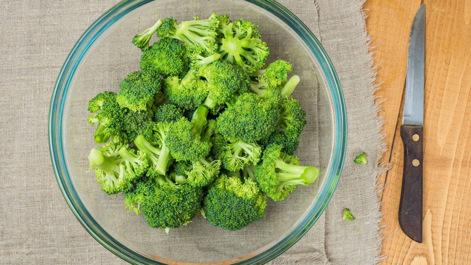 西蘭花盛於透明碗中 壁紙 — 新鮮的綠色西蘭花小朵填滿一個透明玻璃碗，放在有紋理的布上，旁邊有木桌和一把刀。