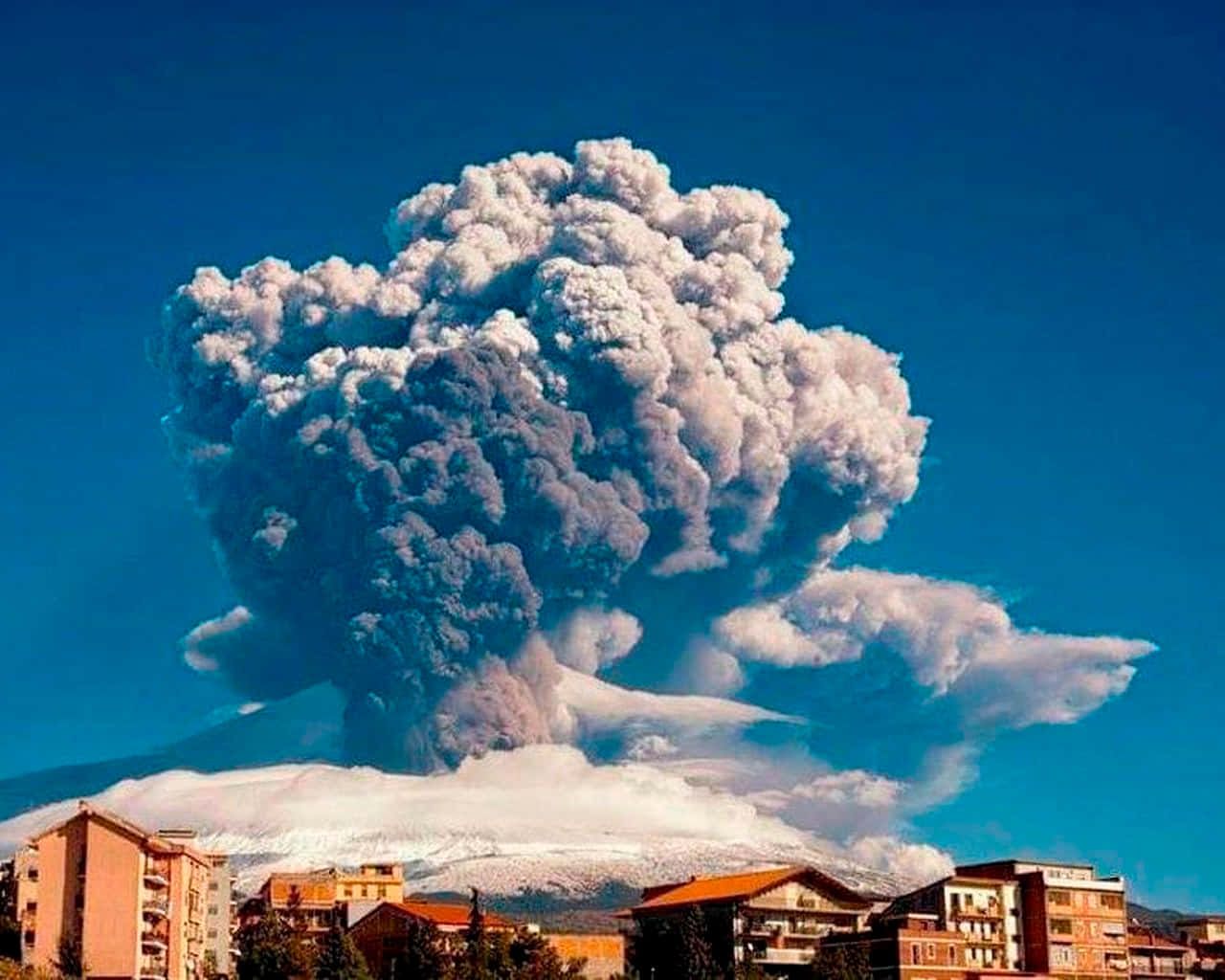 西西里村莊與埃特納火山背景的迷人風景 壁紙 — 大規模火山爆發將巨大的火山灰柱噴向蔚藍天空，下方是白雪覆蓋的山峰和附近的城鎮。