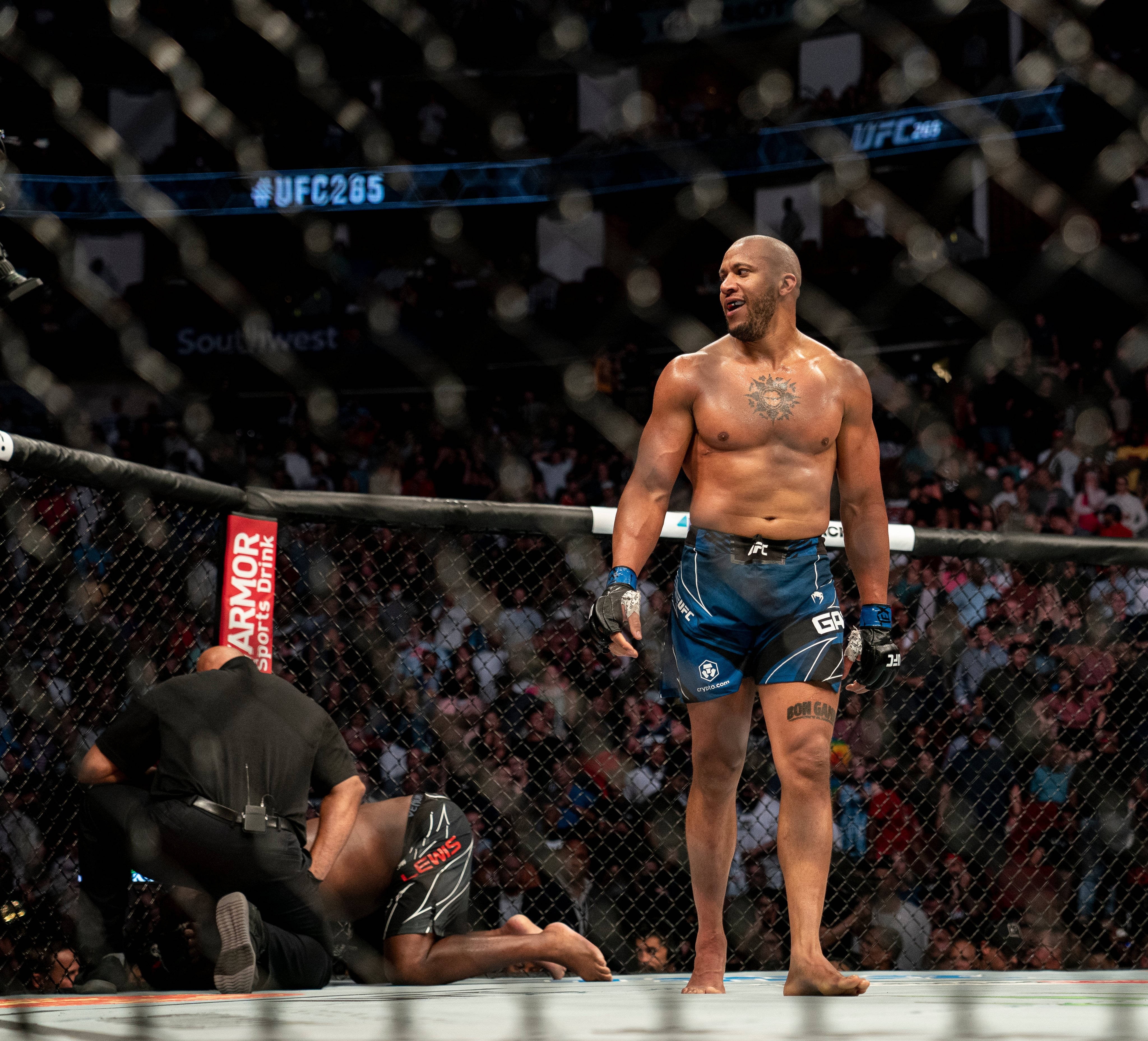 西里爾·甘尼行走 壁紙 — 一位勝利的UFC拳手在八角籠中威風凜凜地站立，而他的對手正由裁判照料。