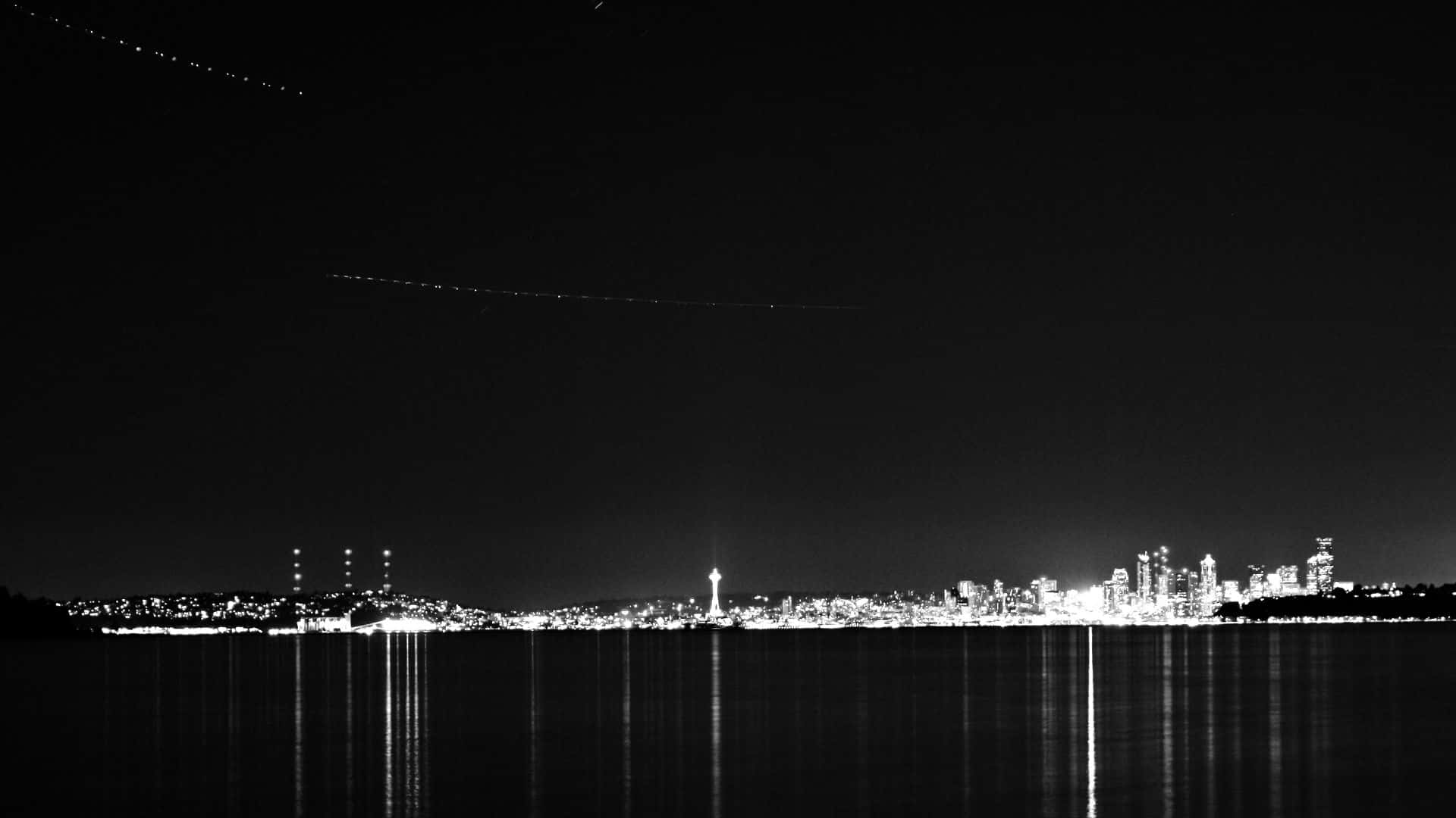 西雅圖天際線黑白夜景 壁紙 — 黑白夜景，城市天際線倒映在平靜的水面上，飛機尾跡劃過漆黑的天空。