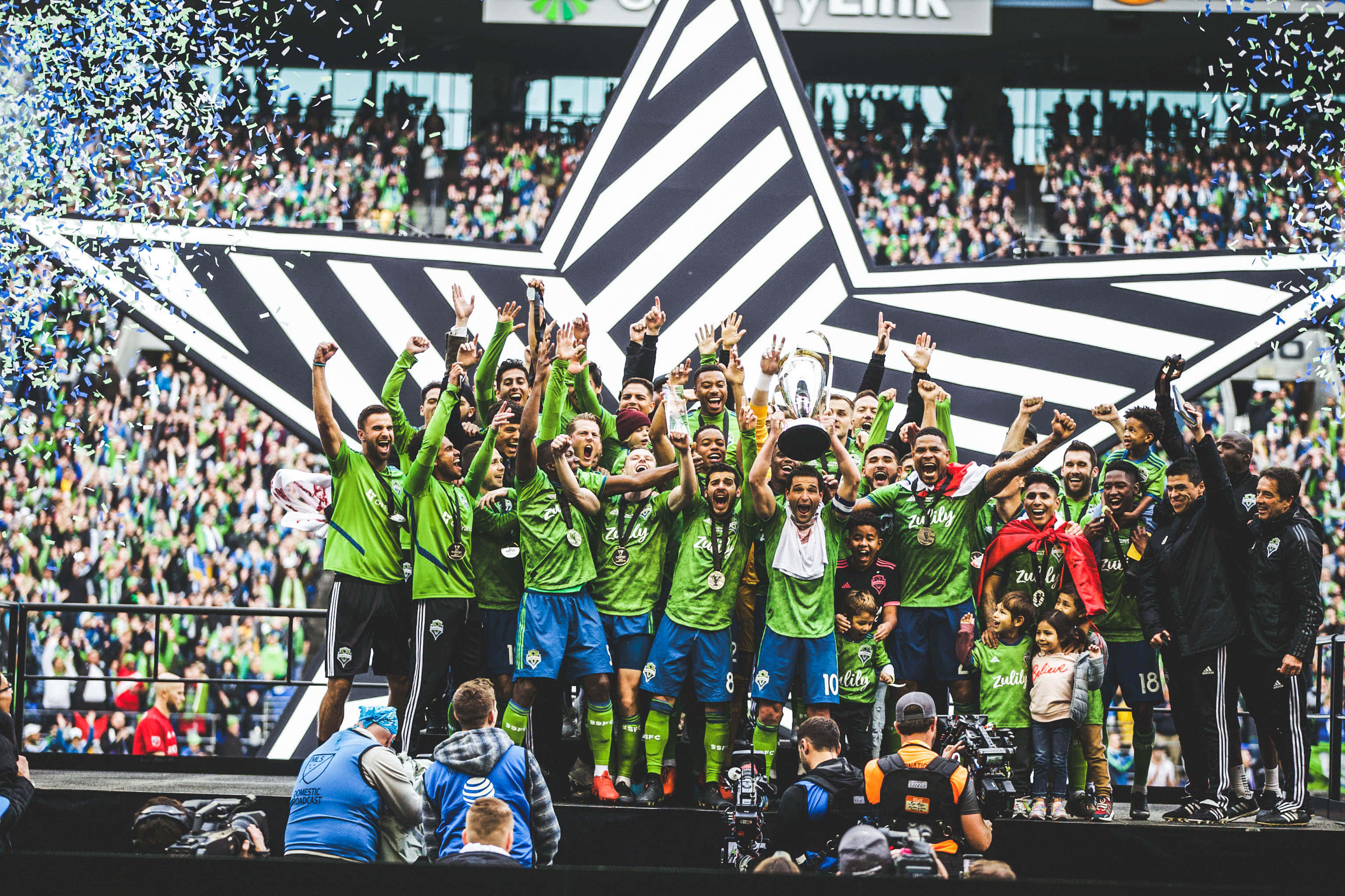 西雅圖音速隊 2019 年 MLS 冠軍慶祝時刻 壁紙 — 一支穿著綠色球衣的足球隊在舞台上慶祝勝利，彩紙飄落，球迷在背景中歡呼。