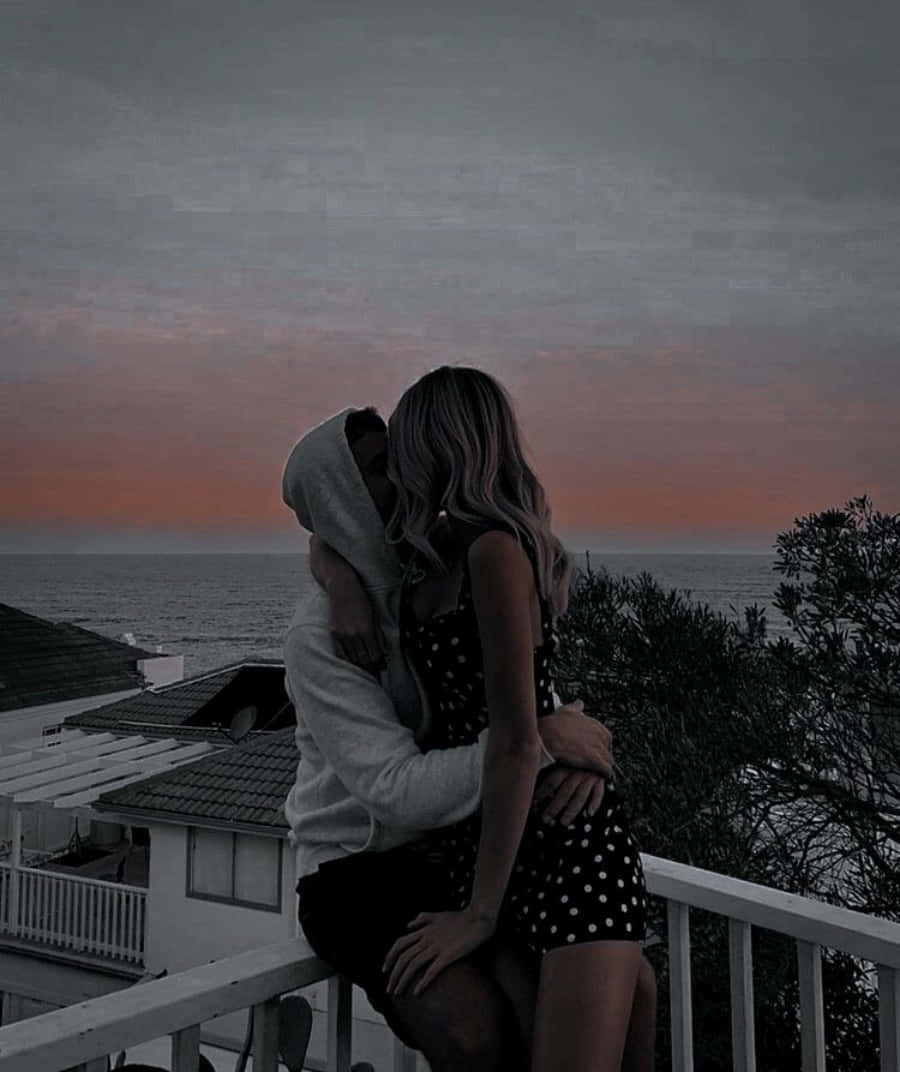 親吻擁抱照片 壁紙 — 一對情侶在陽台上擁抱，而憂鬱的夕陽在海洋上空以柔和的粉紅色和灰色色調塗抹天空。