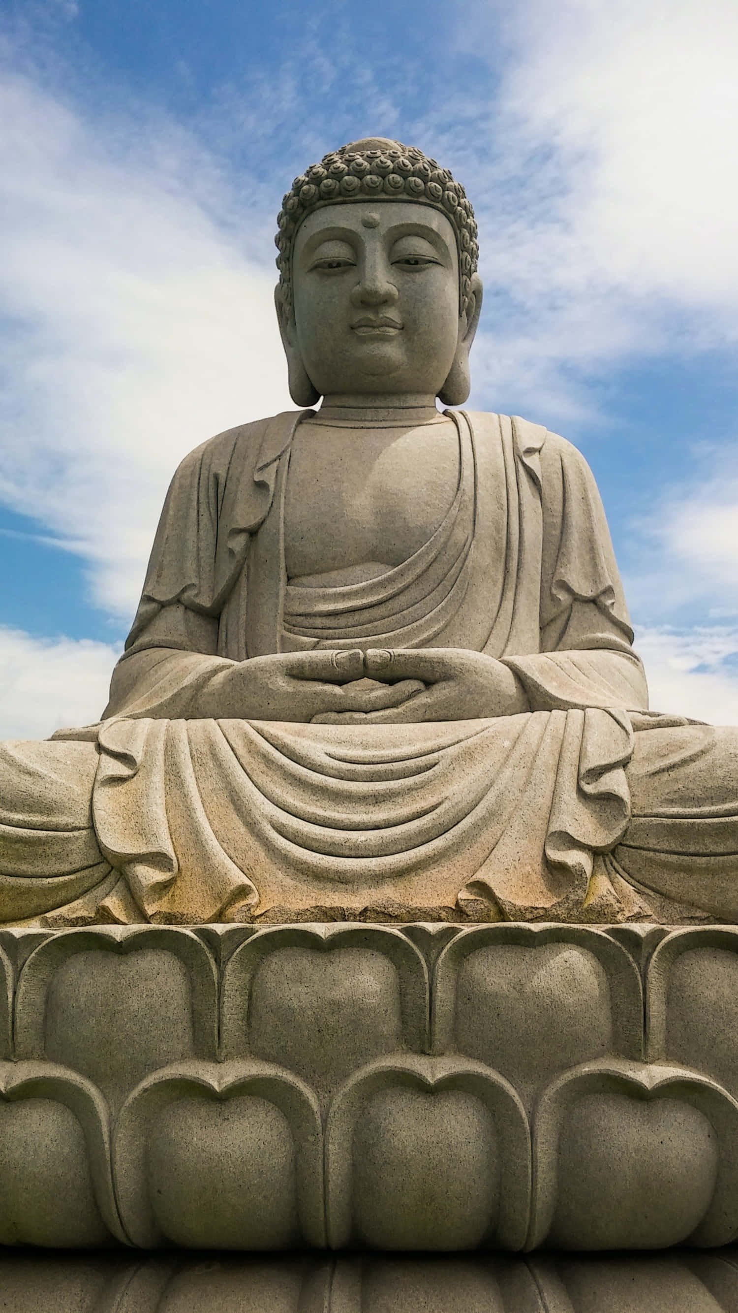 覺醒導師—釋迦牟尼佛 壁紙 — 一尊寧靜的石佛像在藍天白雲下盤腿坐在蓮花座上。