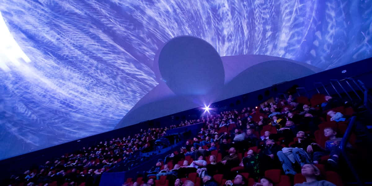 Planetarium 桌布 壁紙 — 觀眾坐在圓頂劇院觀看旋轉藍色星雲和恆星的宇宙投影。