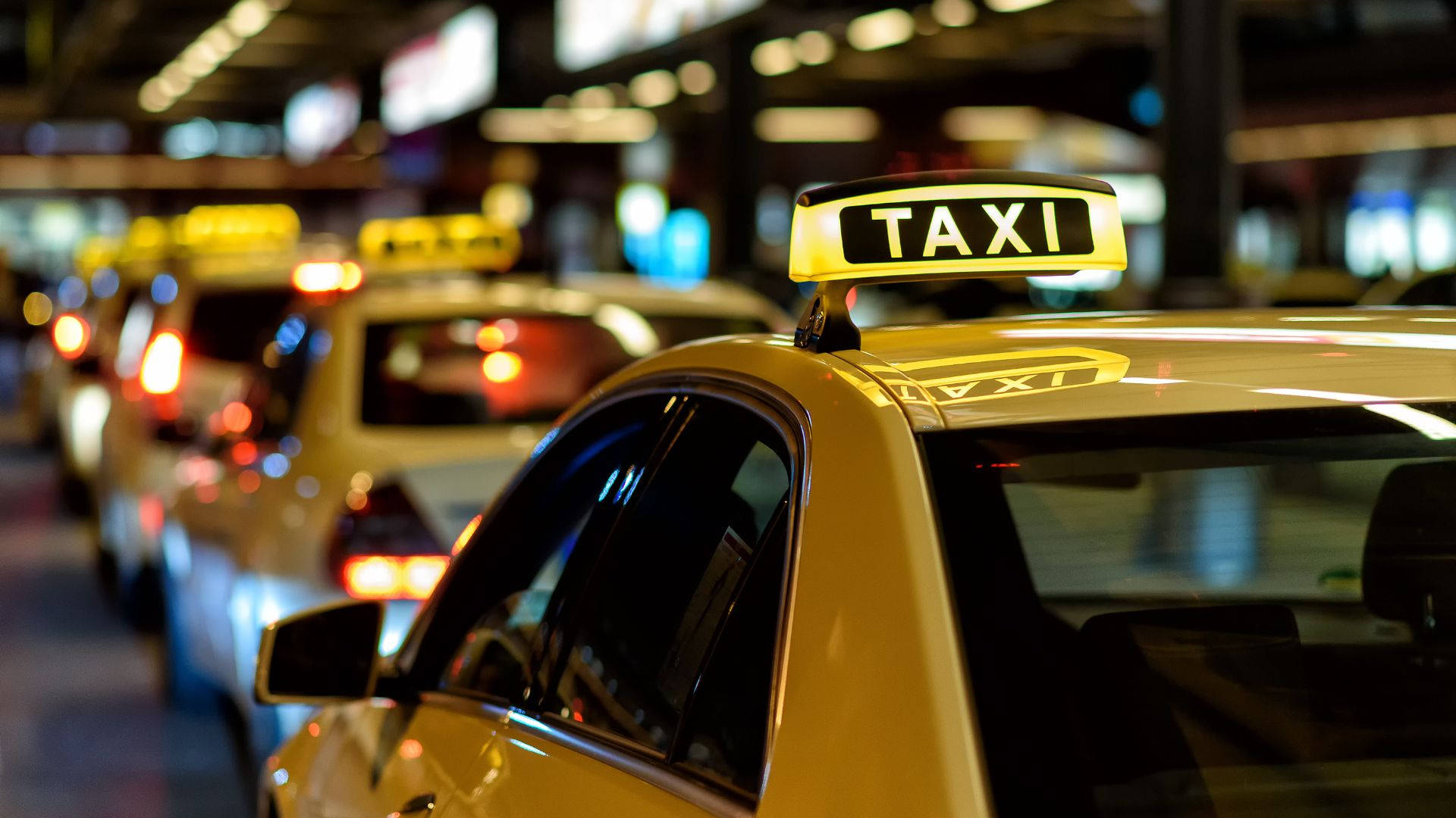 計程車選擇性對焦 壁紙 — 一排黃色的士在夜晚排列，帶有發光的「TAXI」標誌和模糊的城市燈光作為背景。