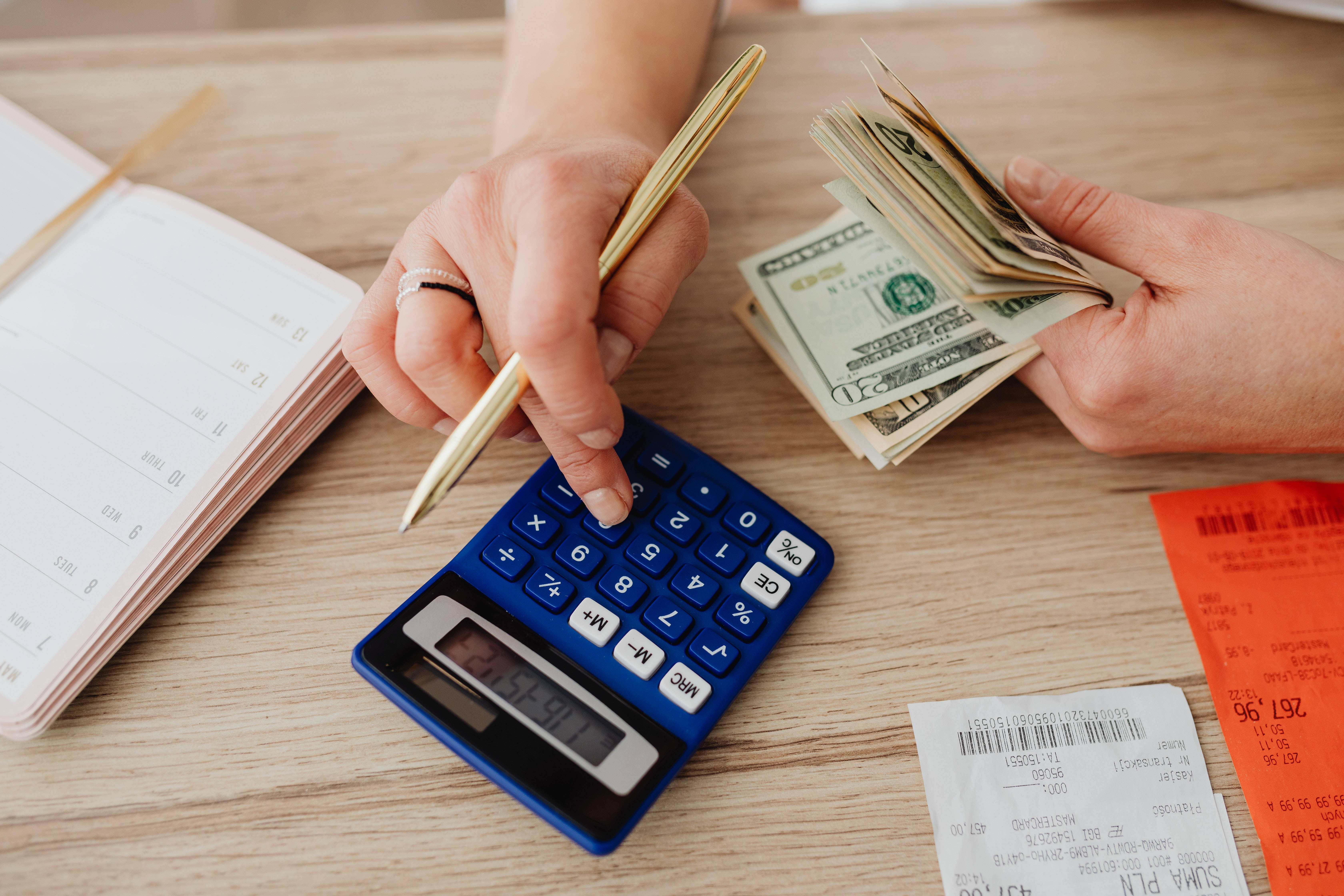 計算通脹金錢 壁紙 — 雙手數現金，並在木桌上使用藍色計算機，旁邊有收據和筆記本。