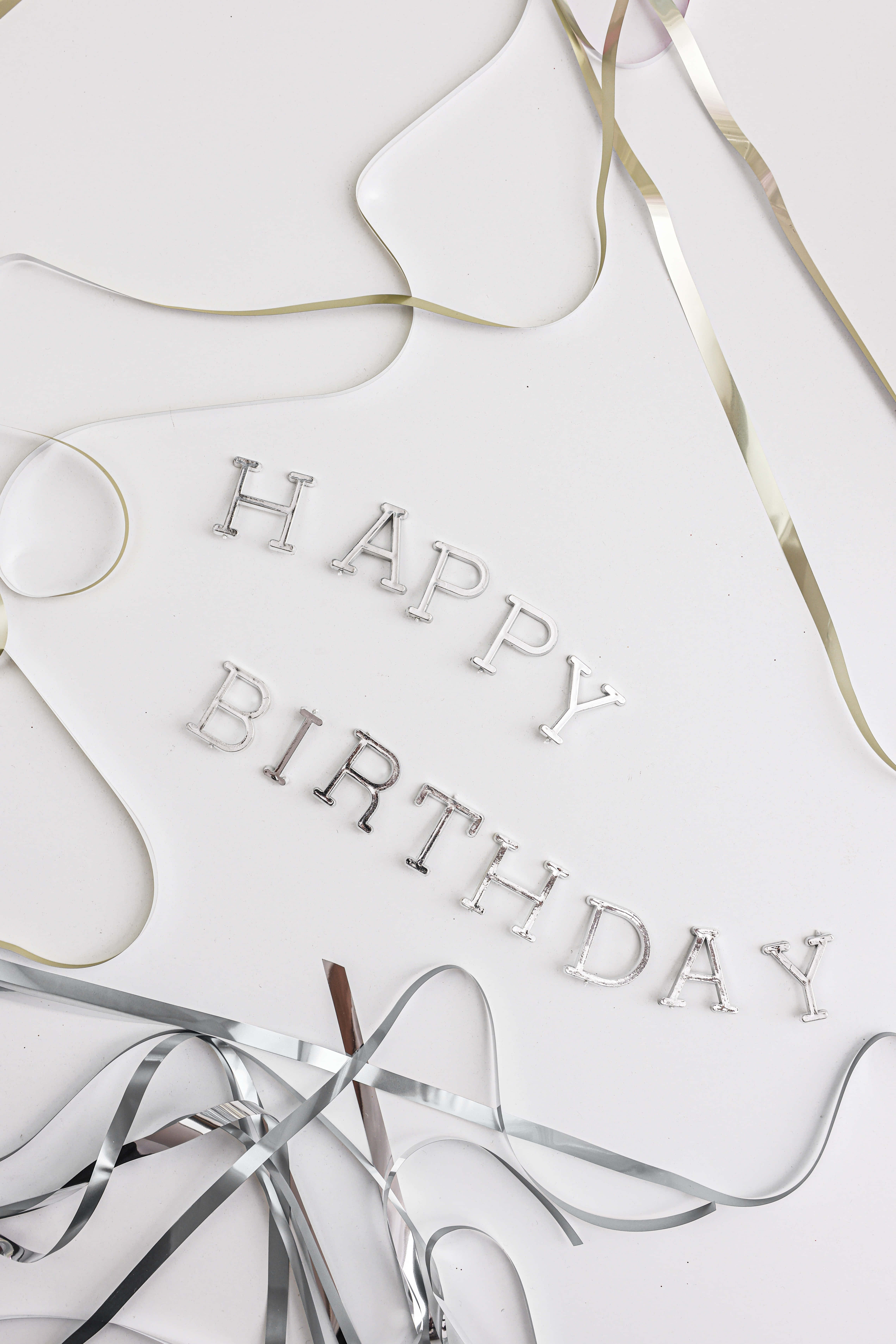 "許個願" 壁紙 — 銀色 'HAPPY BIRTHDAY' 字母和金屬絲帶散落在潔白的表面上。