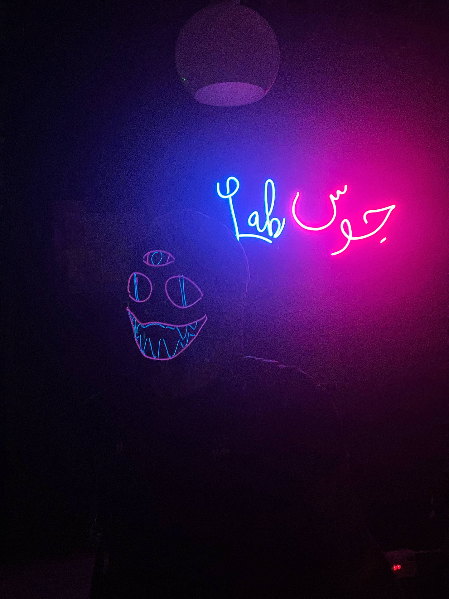 黑暗房間中的人戴著發光的霓虹面具，面具上有 'Lab' 和阿拉伯文字，呈現粉紅色和藍色光。