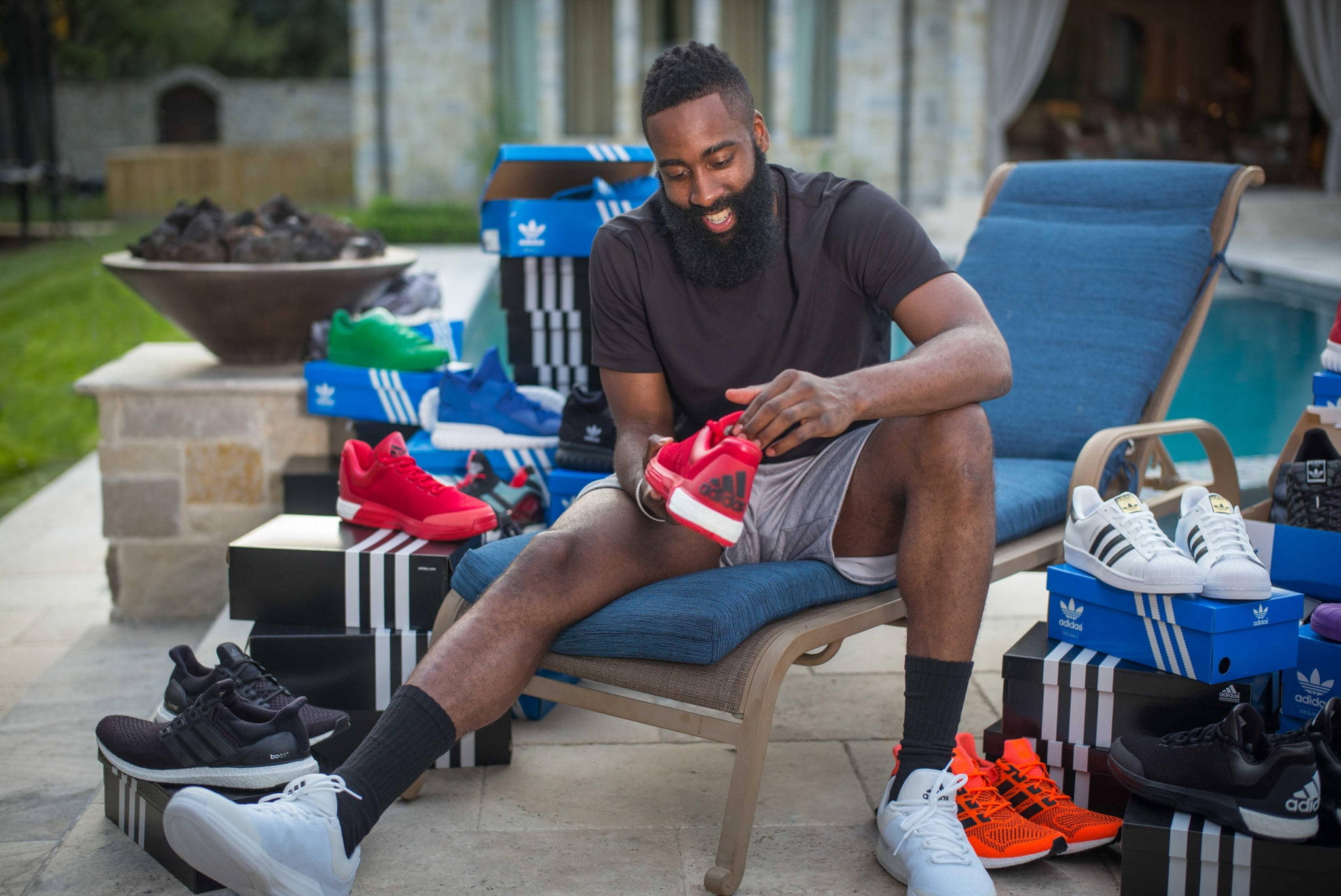 詹姆斯·哈登 Adidas 鞋款系列 壁紙 — 一位留鬍鬚的微笑男子坐在戶外的藍色躺椅上，周圍環繞著色彩繽紛的Adidas運動鞋和盒子。