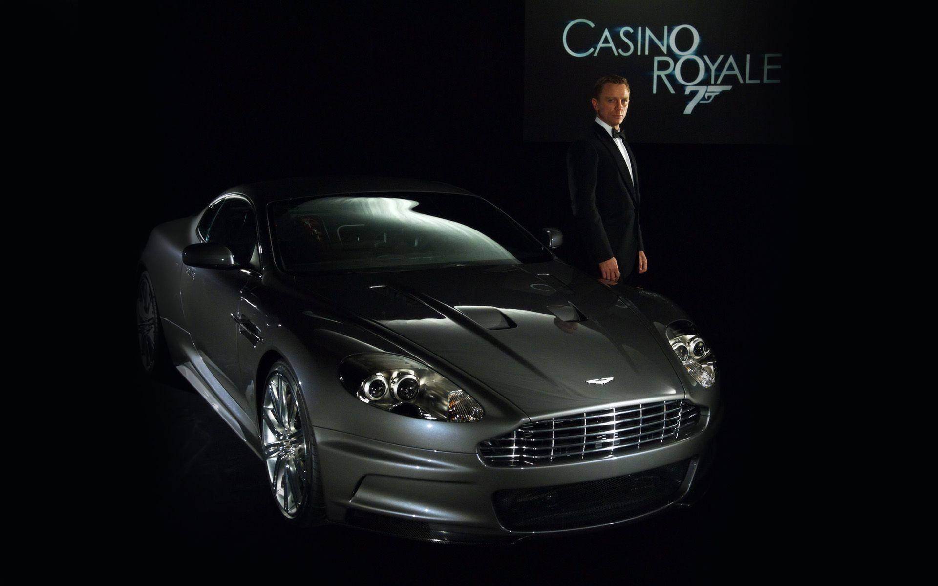 詹姆斯邦德與跑車 壁紙 — 一輛時尚的銀色Aston Martin DBS在聚光燈下閃閃發光，丹尼爾·克雷格站在旁邊，背景是黑色，帶有Casino Royale標誌。