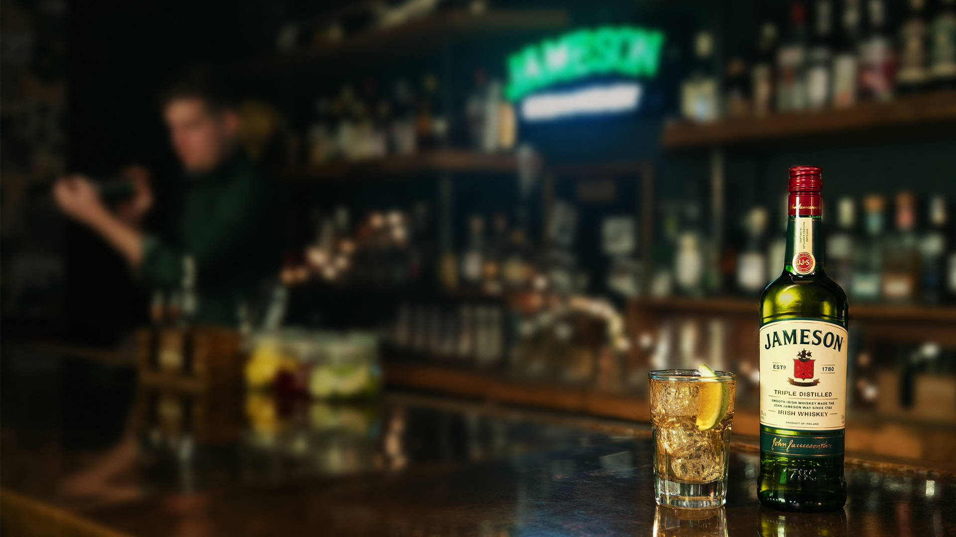 詹姆森酒吧與飲品 壁紙 — 一瓶Jameson愛爾蘭威士忌和一杯加冰和檸檬的玻璃杯放在拋光的吧檯上，背景中調酒師模糊，環境燈光溫暖。
