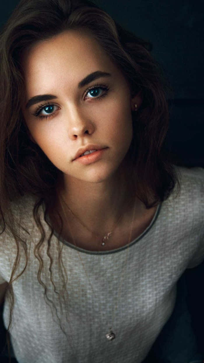 認真的青春少女 壁紙 — 一位年輕女性，擁有引人注目的藍色眼睛和波浪狀棕色頭髮，在柔和光線的肖像中直視鏡頭。