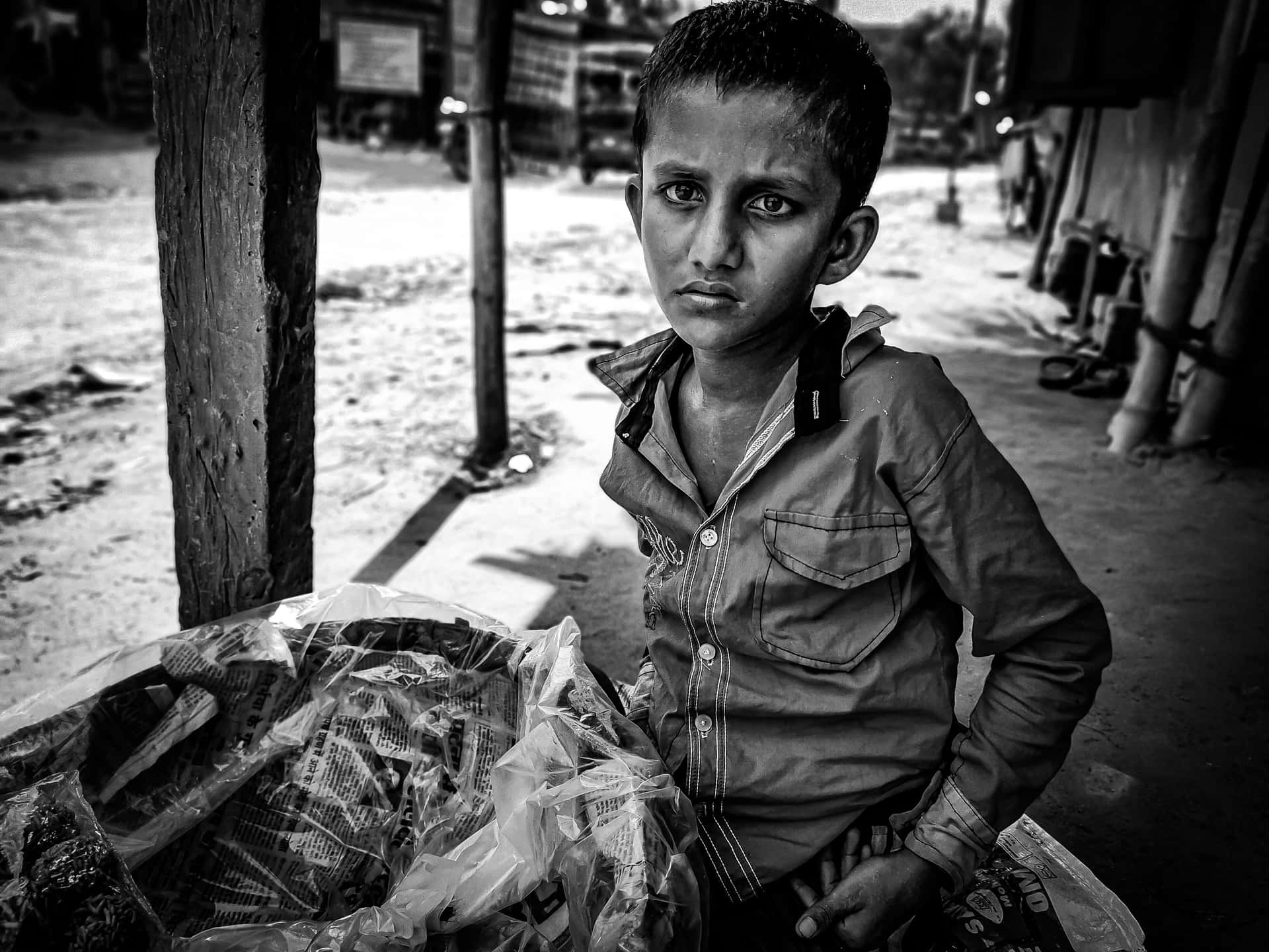 一個穿著鈕扣襯衫的小男孩，在鄉村戶外環境中，旁邊有塑膠袋，直視鏡頭。