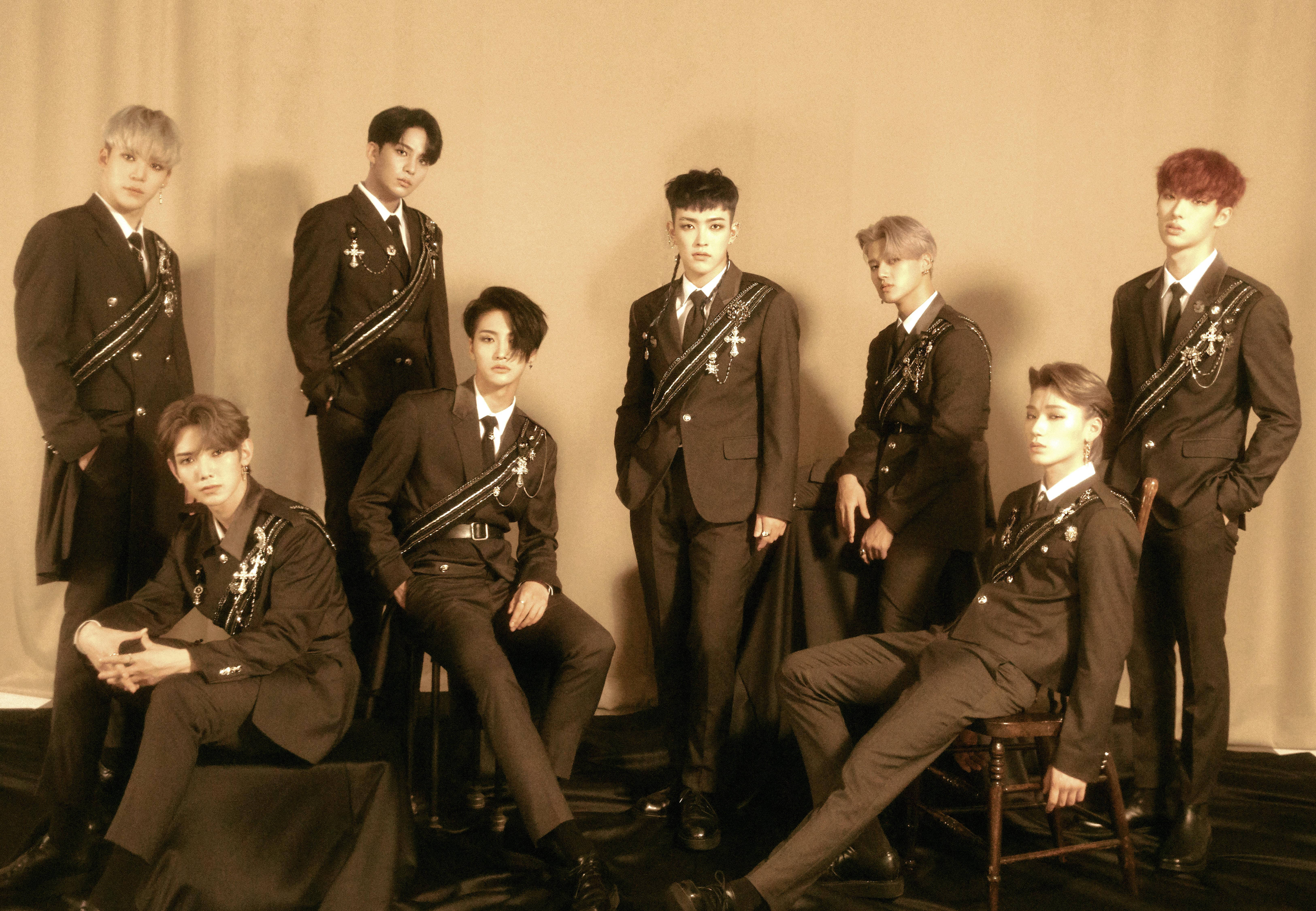 認識ATEEZ，來自韓國的動感K-pop男子團體 壁紙 — 七名身穿華麗深色西裝的男子在溫暖的棕褐色燈光下，自信地坐在椅子上或站在樸素的背景前。