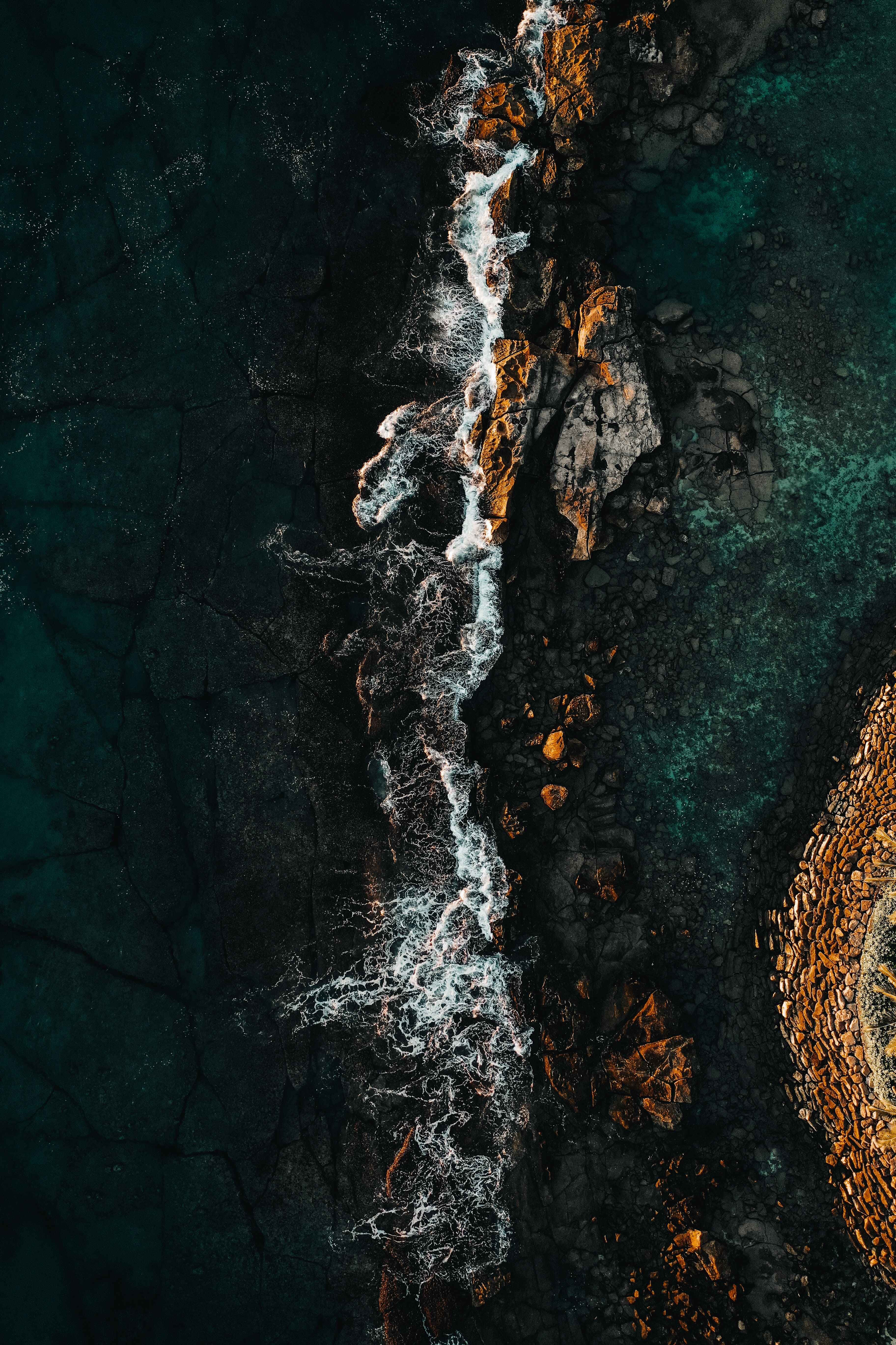 誘人奧阿胡水域 iPhone 背景 壁紙 — 航拍視角，洶湧波浪拍打崎嶇的深色岩石，碧綠海水與金色亮點。