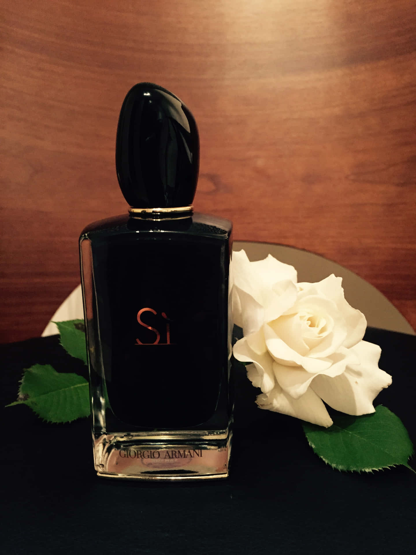 黑色 Giorgio Armani 香水瓶與白玫瑰和綠葉在深色表面上。